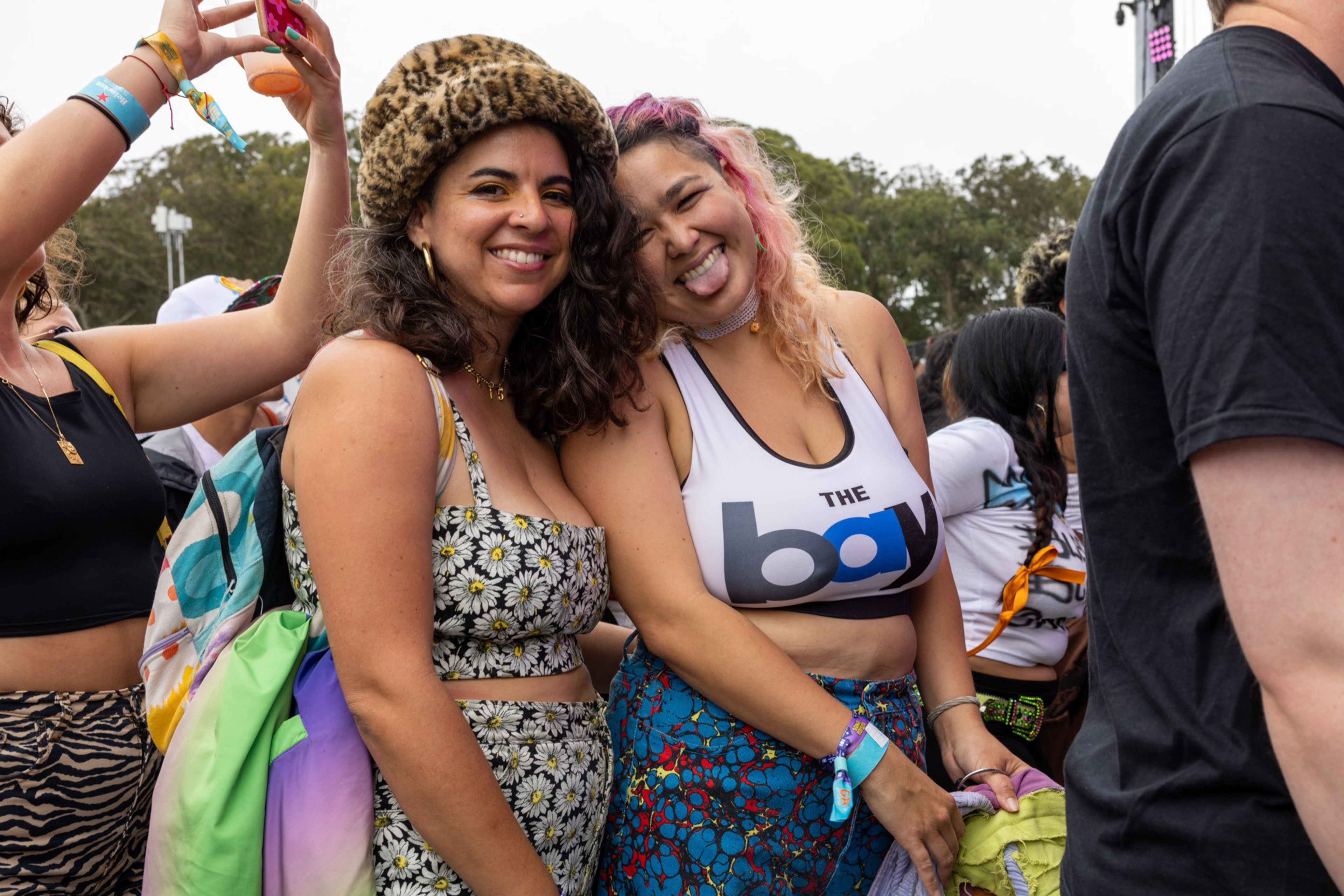 Two women smile and pose at an outdoor event; one wears a leopard print hat and floral outfit, the other a "The Bay" crop top with colorful skirt.