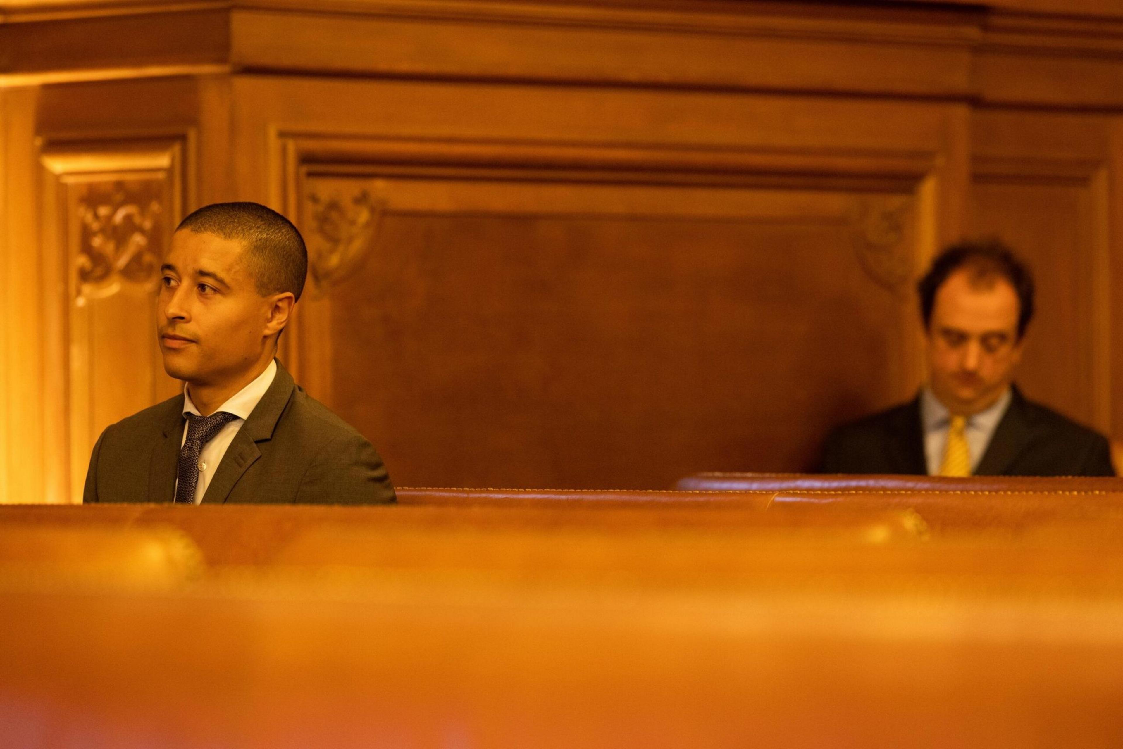 Two men in suits sit separately on wooden benches in a dimly lit room, one looking away thoughtfully and the other looking down.