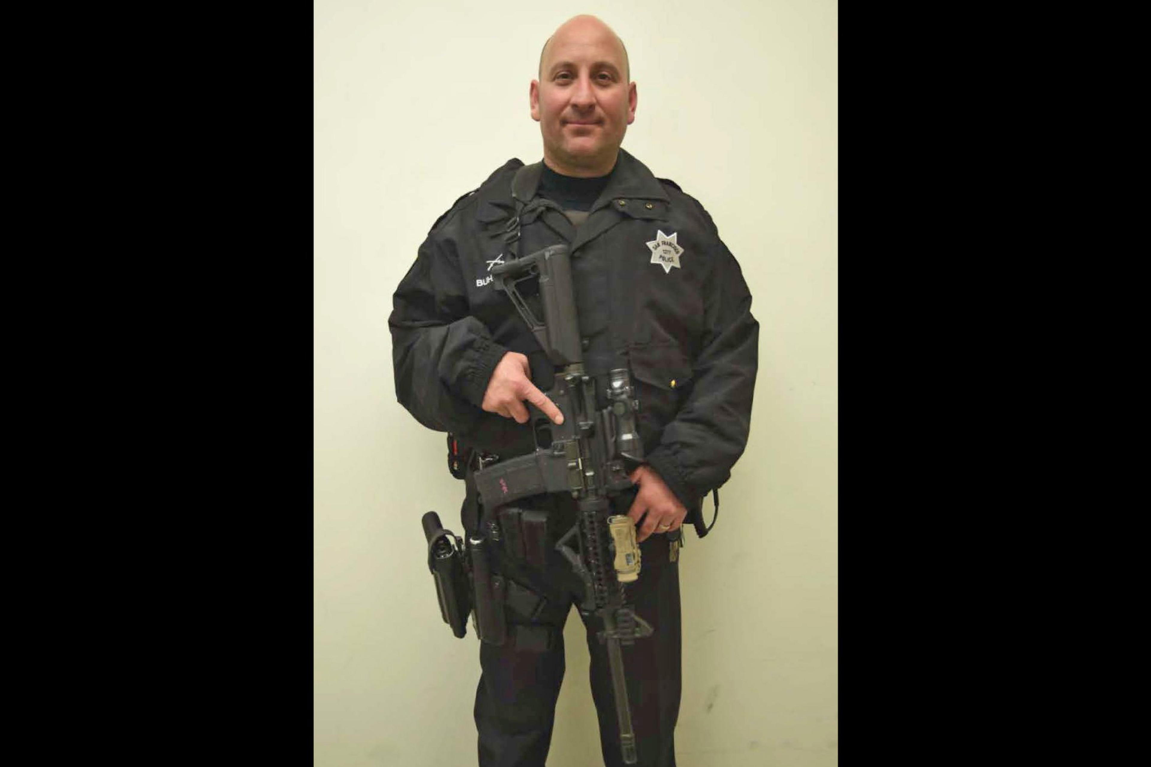 A man in a black police tactical uniform stands against a plain wall, holding a large rifle in front of him with both hands.