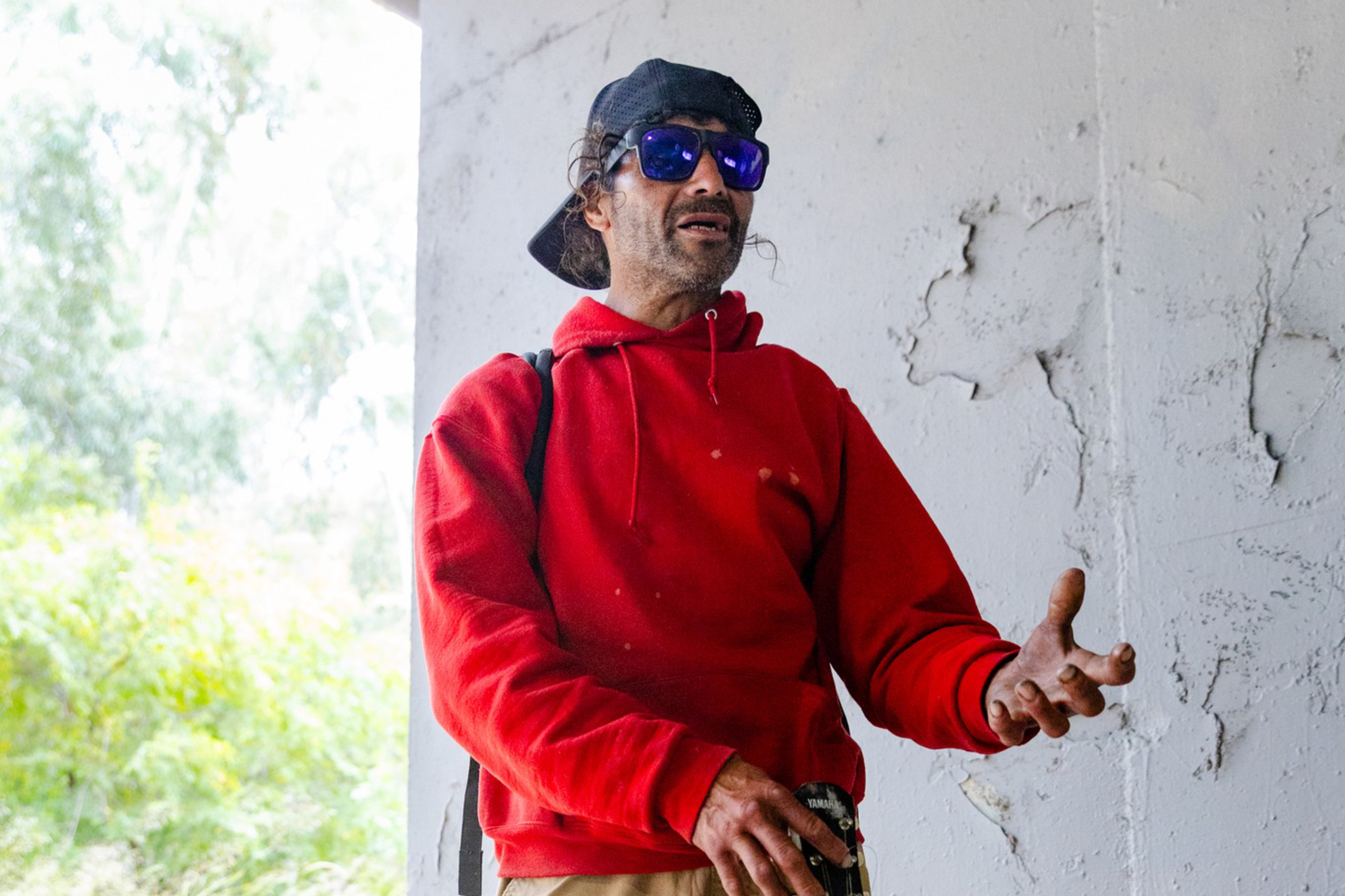 A man in a red hoodie, beige pants, and dark boots holds a black guitar with a red strap, wearing a black cap backward and blue sunglasses.