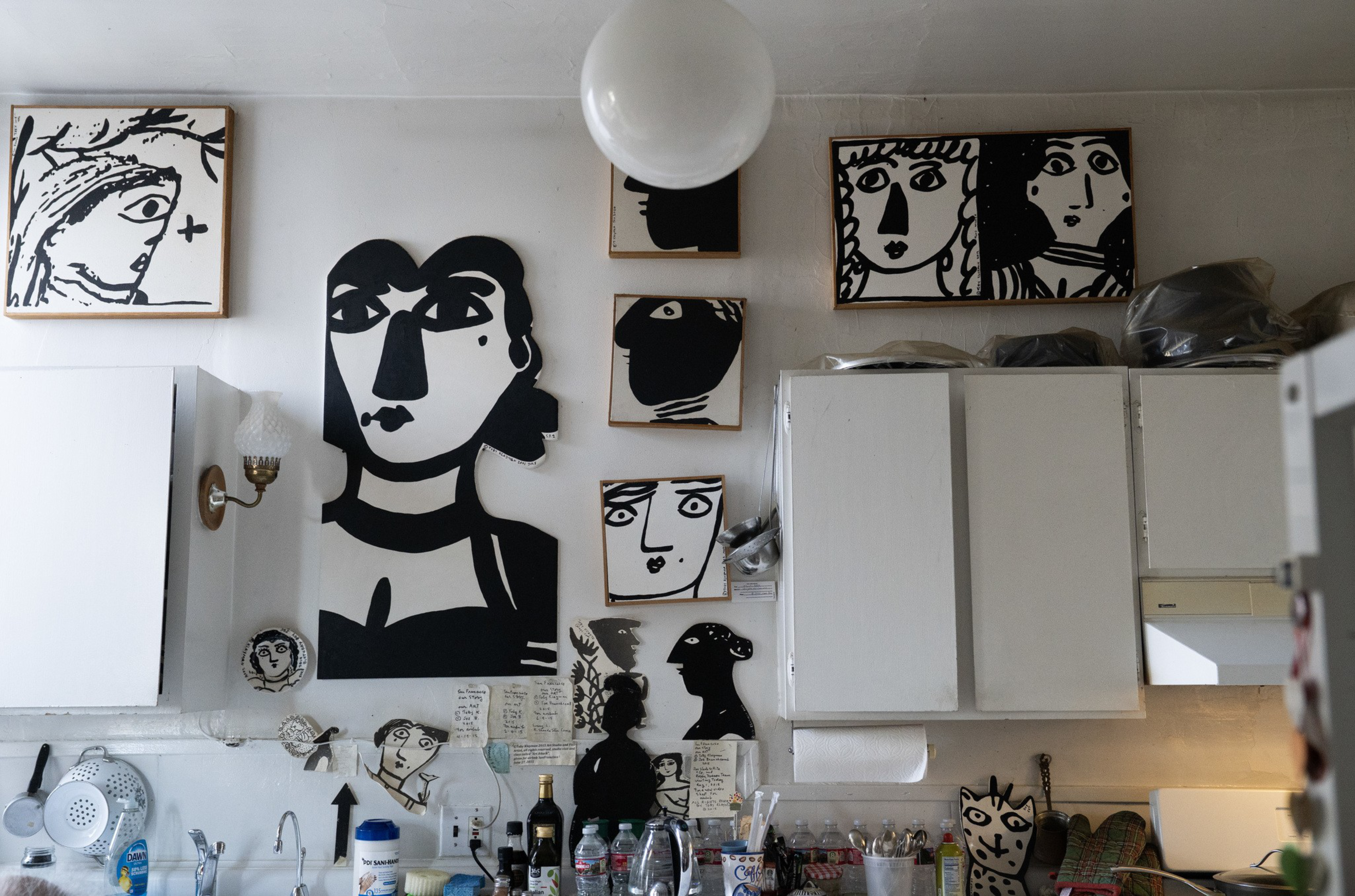 Black-and-white abstract face art decorates a kitchen wall above white cabinets and a cluttered countertop with bottles and utensils.