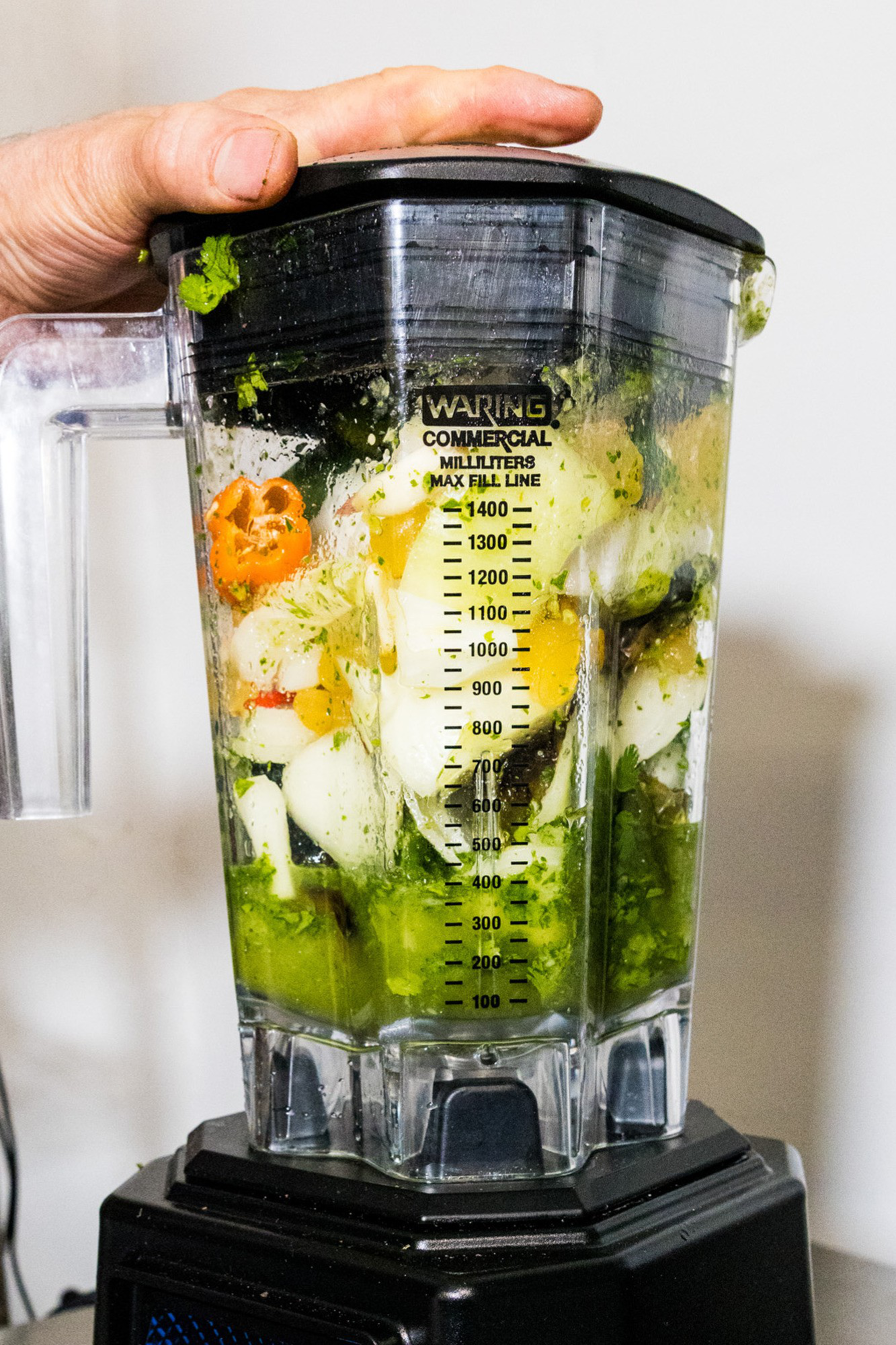 A blender filled with cut vegetables, including peppers, onions, and herbs. A hand presses down on the lid, ready for blending.