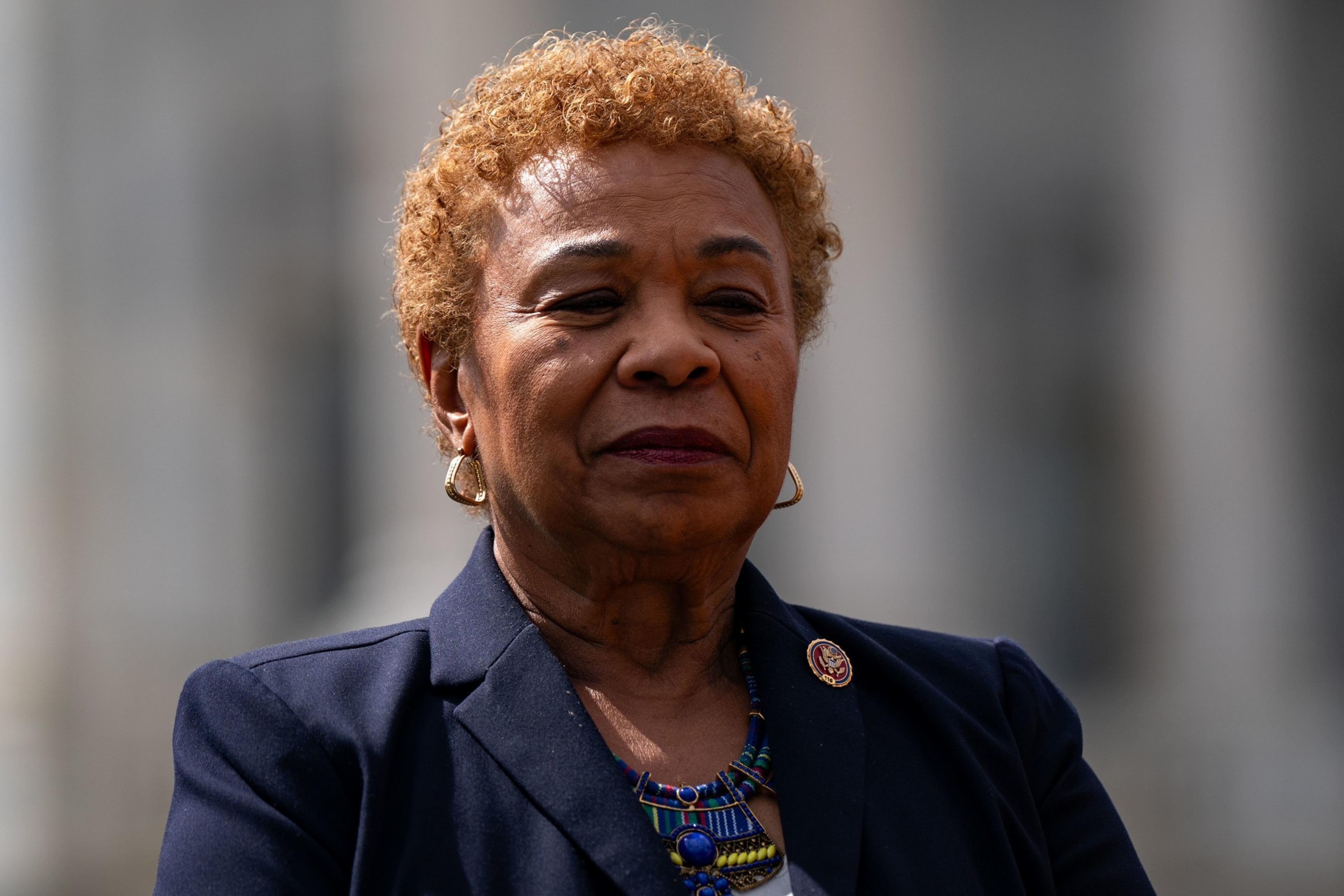 A person with short curly hair is wearing a navy blazer and colorful beaded necklace, with a pin on the lapel. The background is blurred.