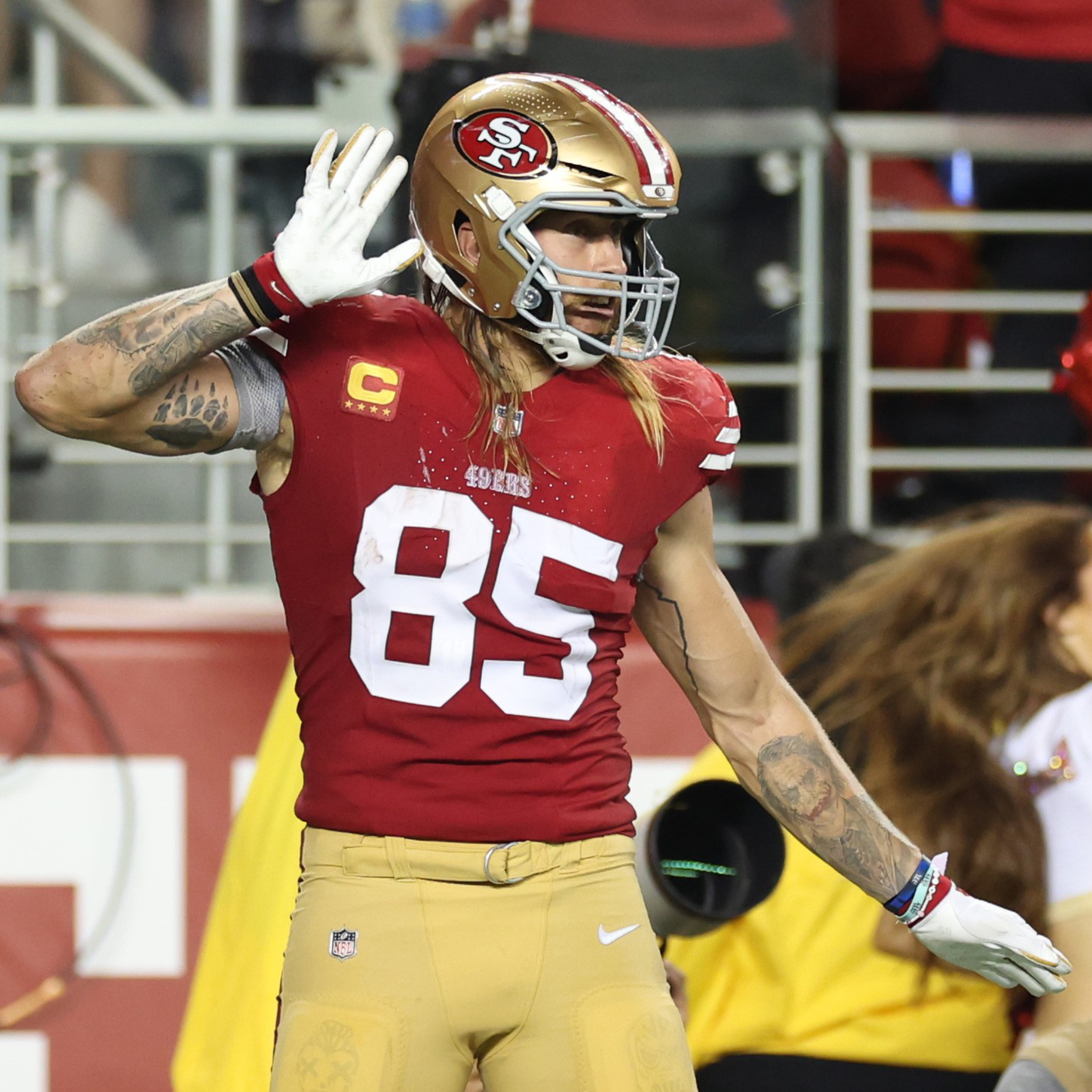 A football player in a red and gold uniform with the number 85, wearing a helmet, holds his hand to his ear on the field. A cheerleader is in the background.