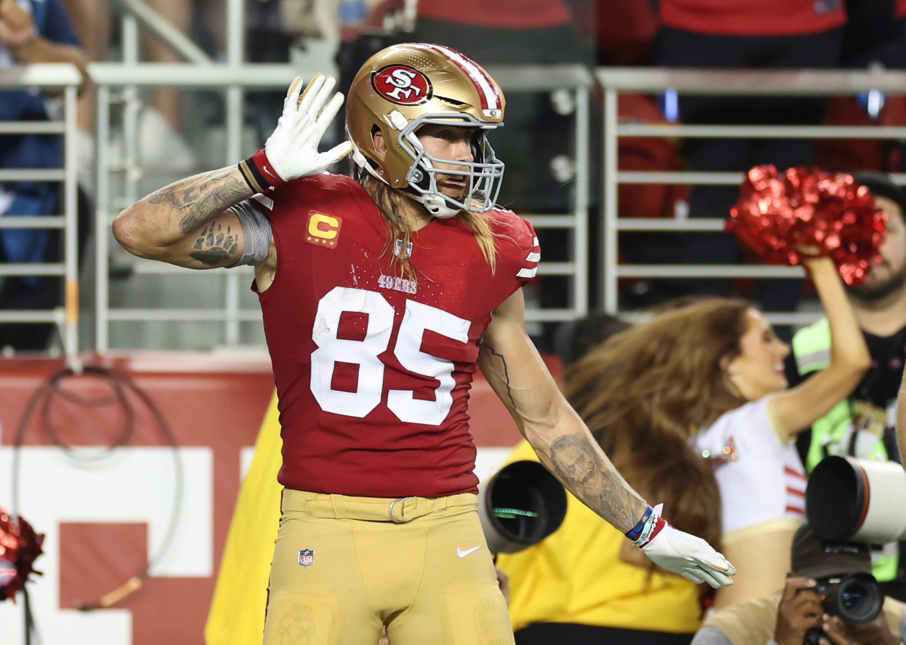 A football player in a red and gold uniform with the number 85, wearing a helmet, holds his hand to his ear on the field. A cheerleader is in the background.