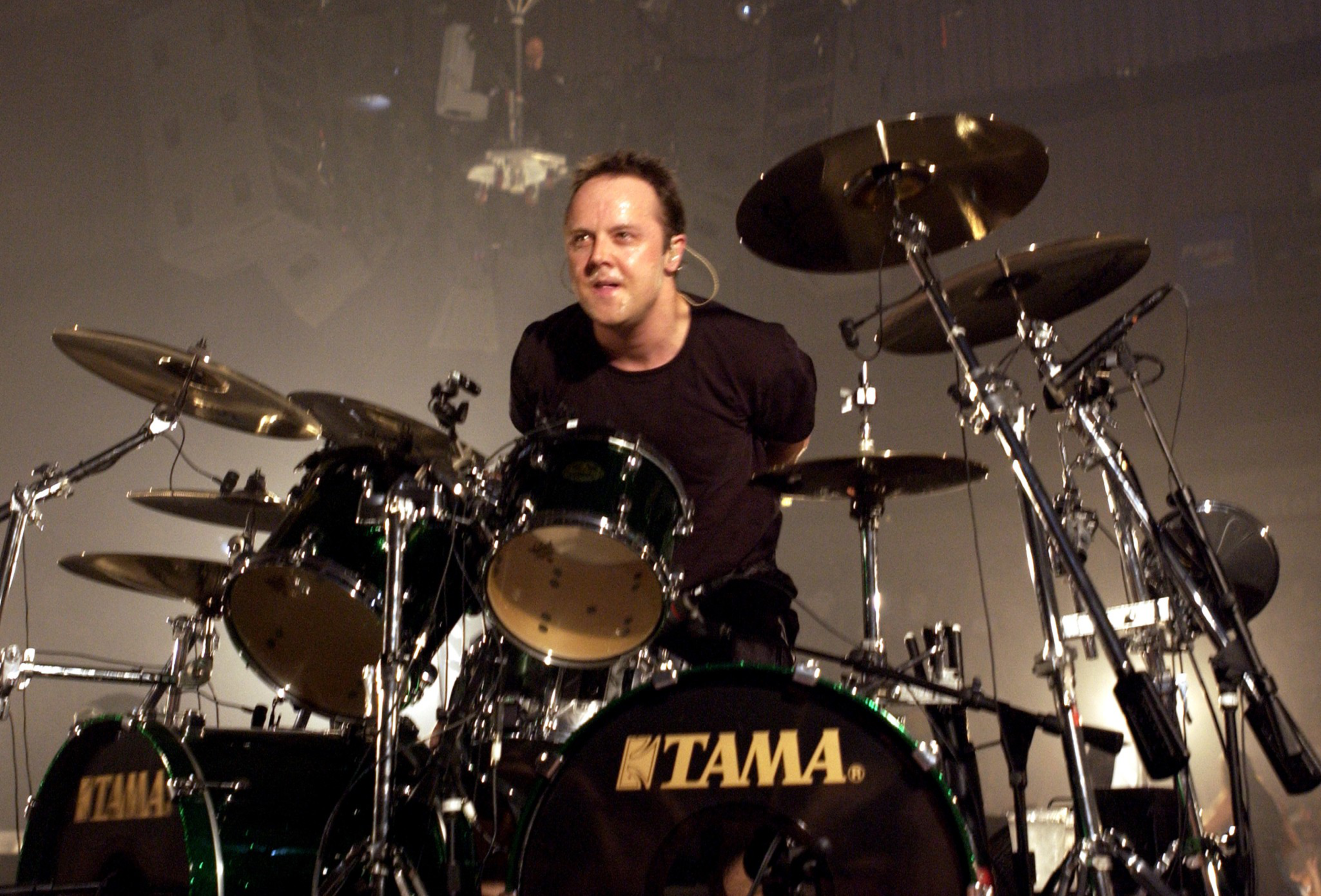 A drummer sits focused behind a Tama drum set with cymbals, mid-performance under stage lights.