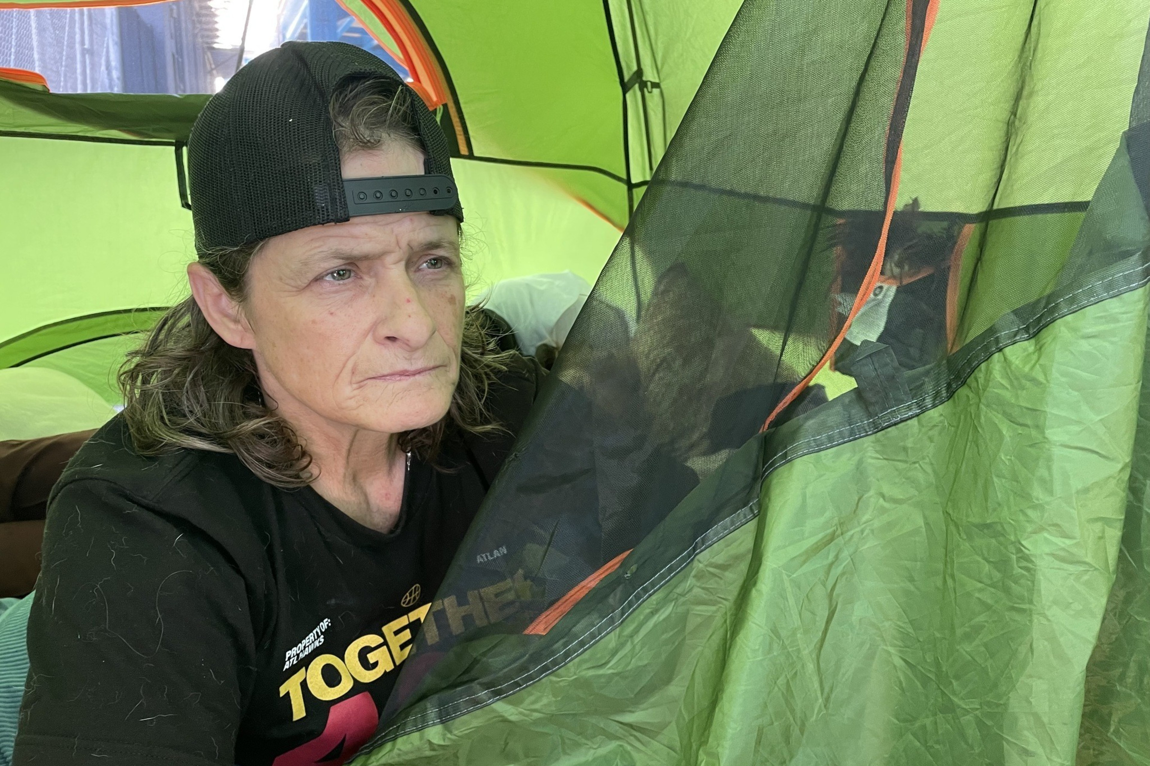 A person with long hair wearing a backwards cap sits inside a green tent, holding the tent flap. The person has a thoughtful expression and a cross tattoo on their arm.