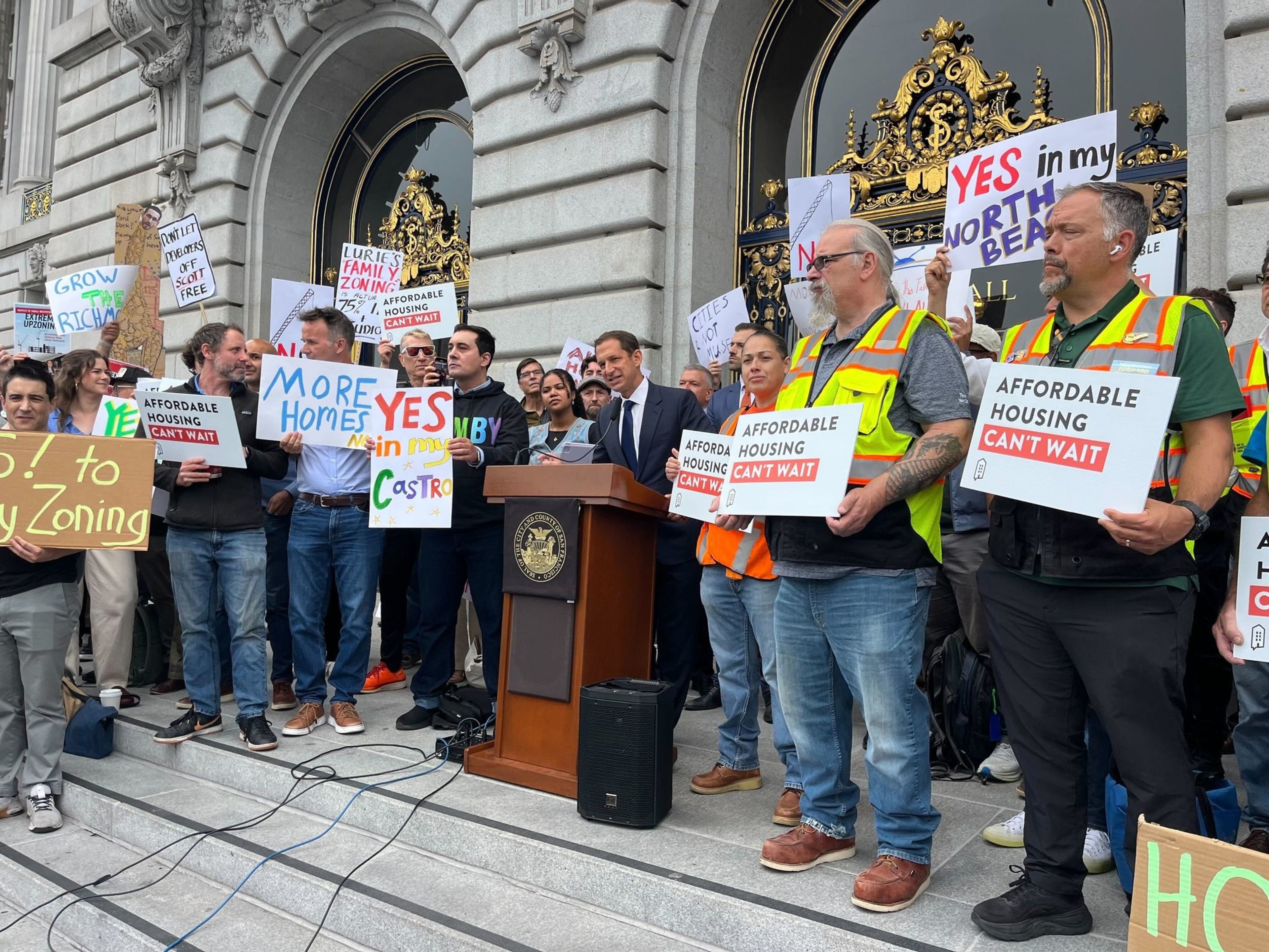 A diverse crowd holds signs advocating for affordable housing and more homes, while a speaker addresses them from a podium on grand building steps.