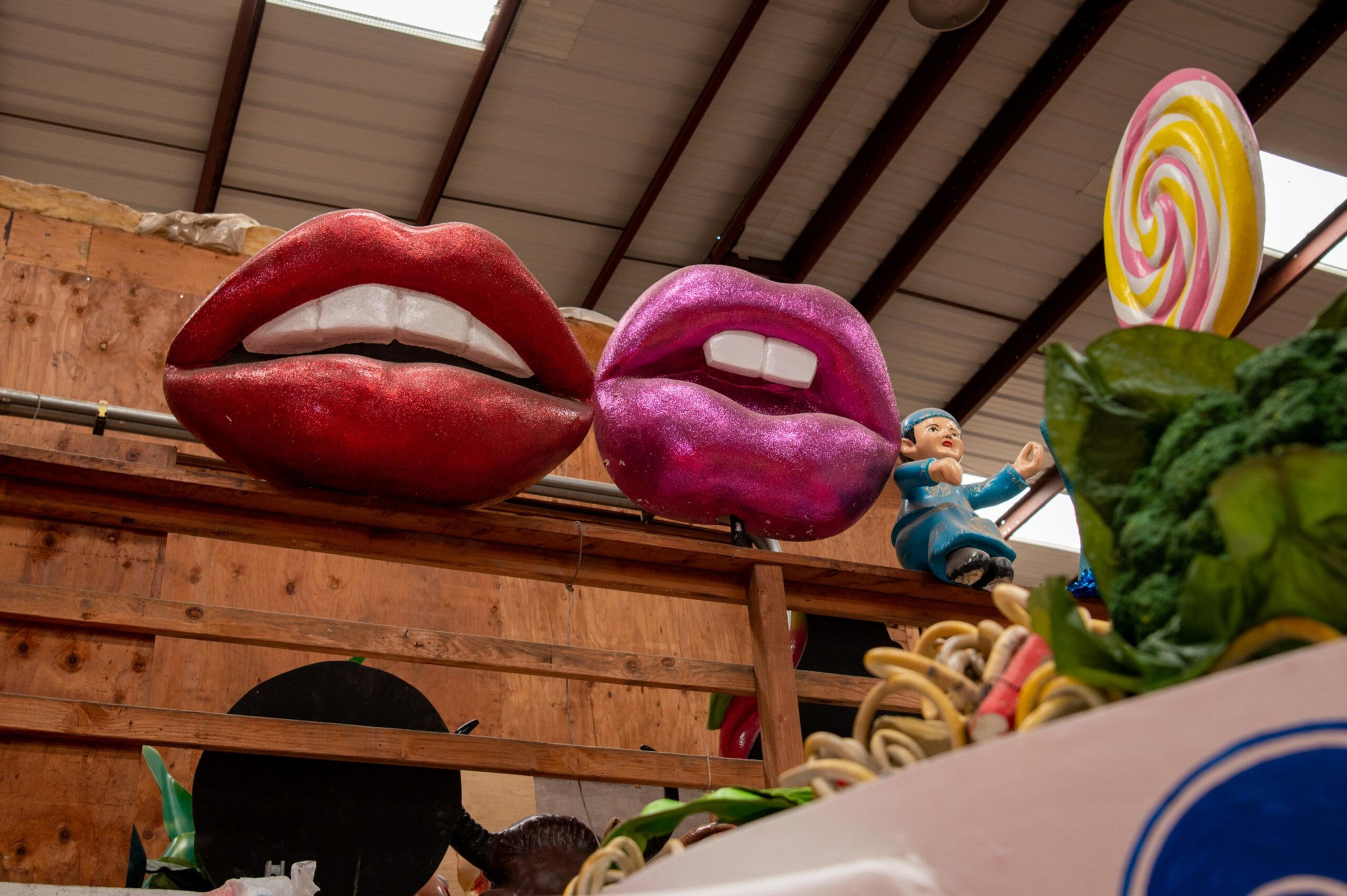 Two large, colorful lips sculptures—one red and one purple—are mounted on a wooden wall inside a warehouse with various props visible below.