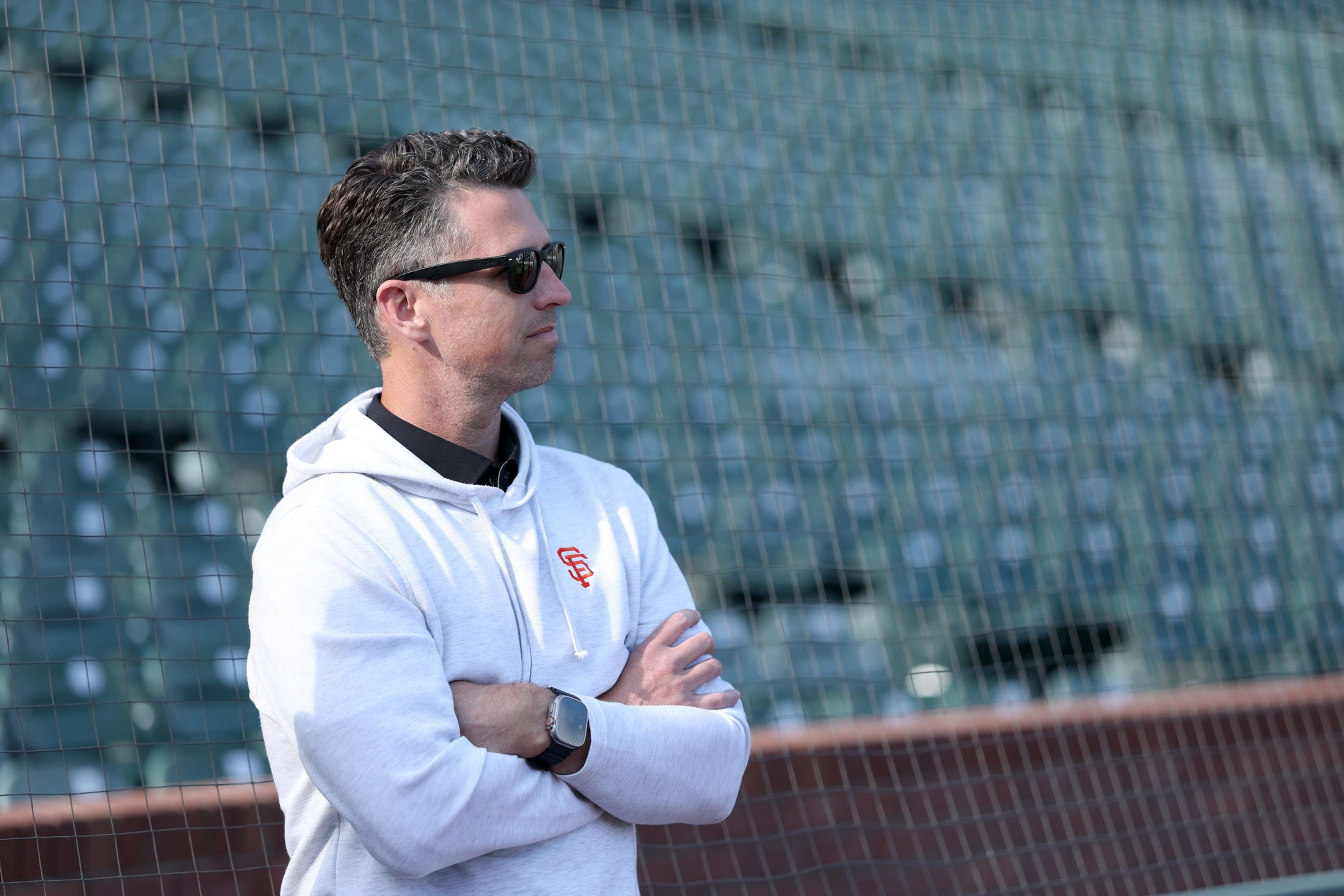 A man wearing sunglasses and a white hoodie with a logo stands with arms crossed, looking to the side in front of empty stadium seats behind netting.