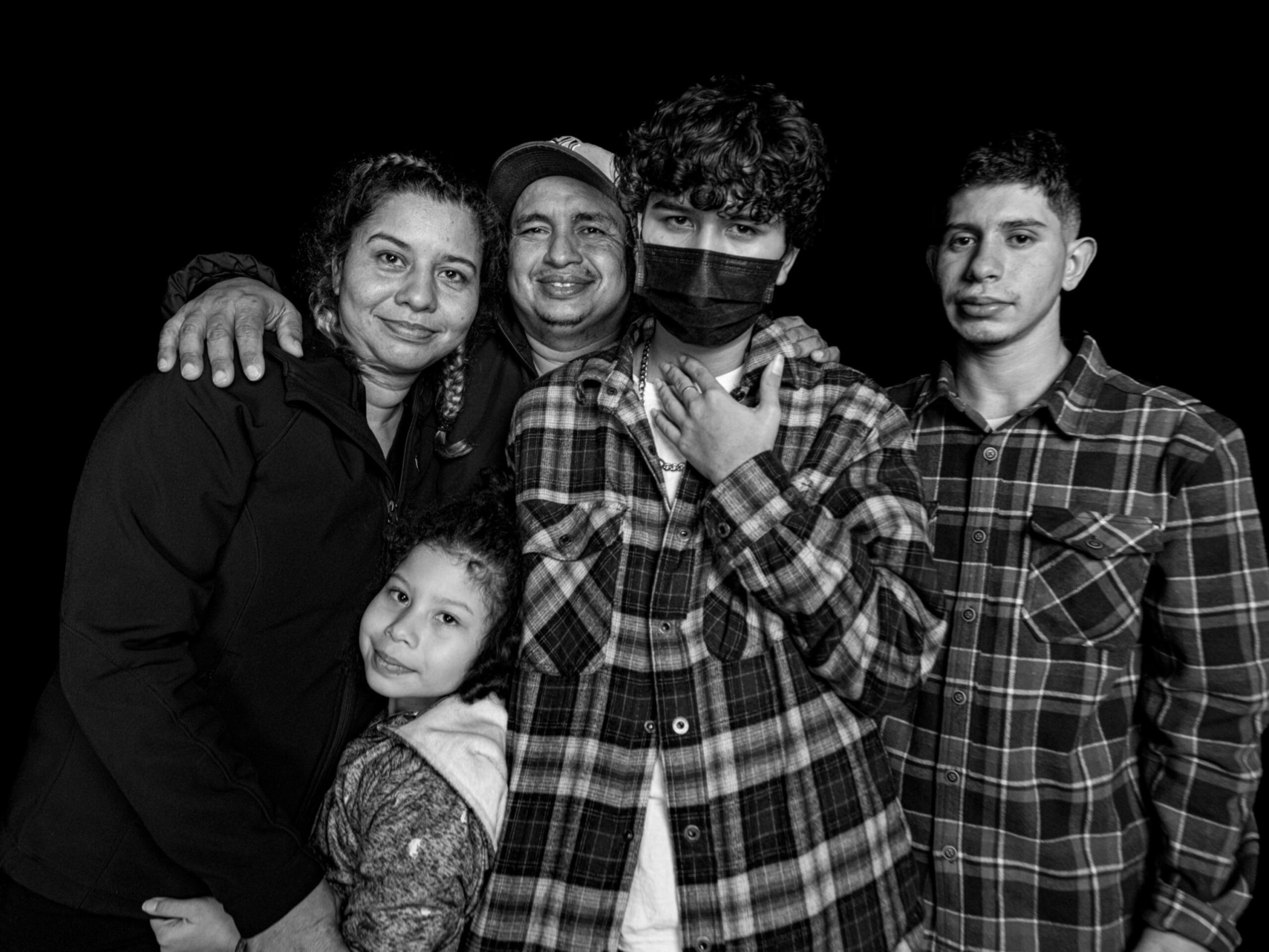 A family of five poses closely, with a woman, man, and child on left smiling, a teen in a mask in the center, and a young man in plaid on the right.