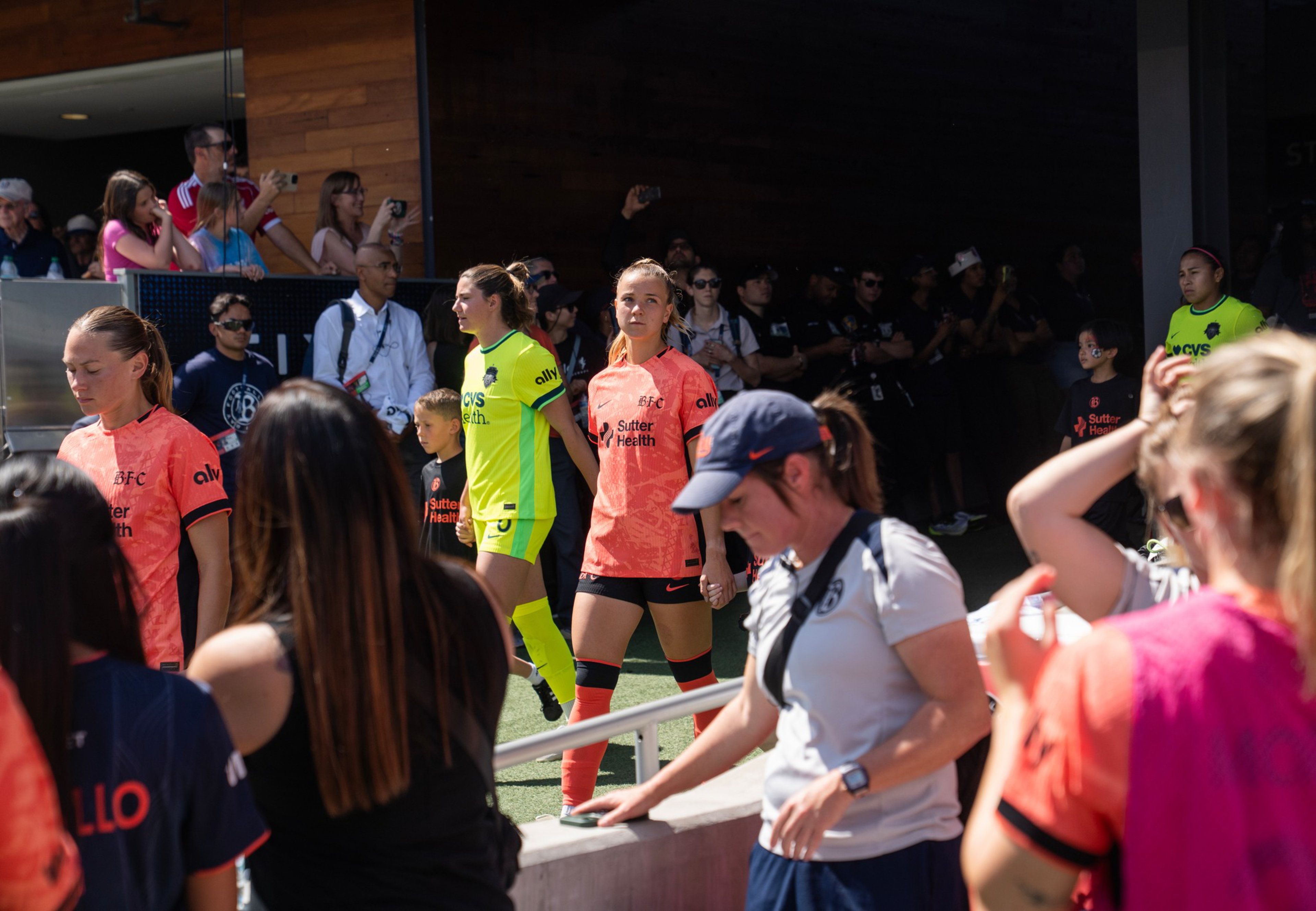 Female soccer players in orange and neon yellow uniforms walk onto the field, surrounded by fans and personnel taking photos and watching.