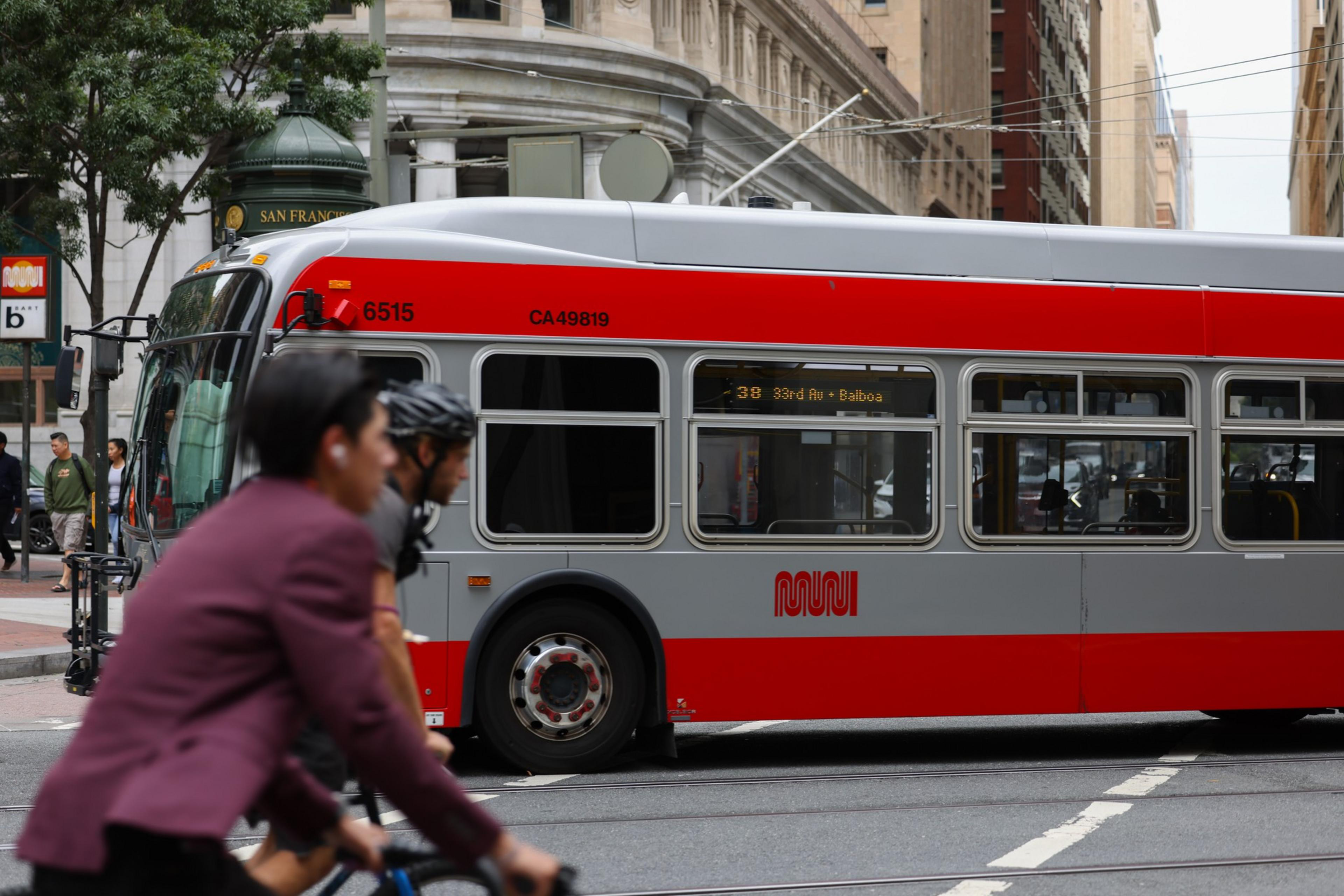 A red and gray city bus marked MUNI and route 38 drives through an urban street while two cyclists pass by in the foreground.