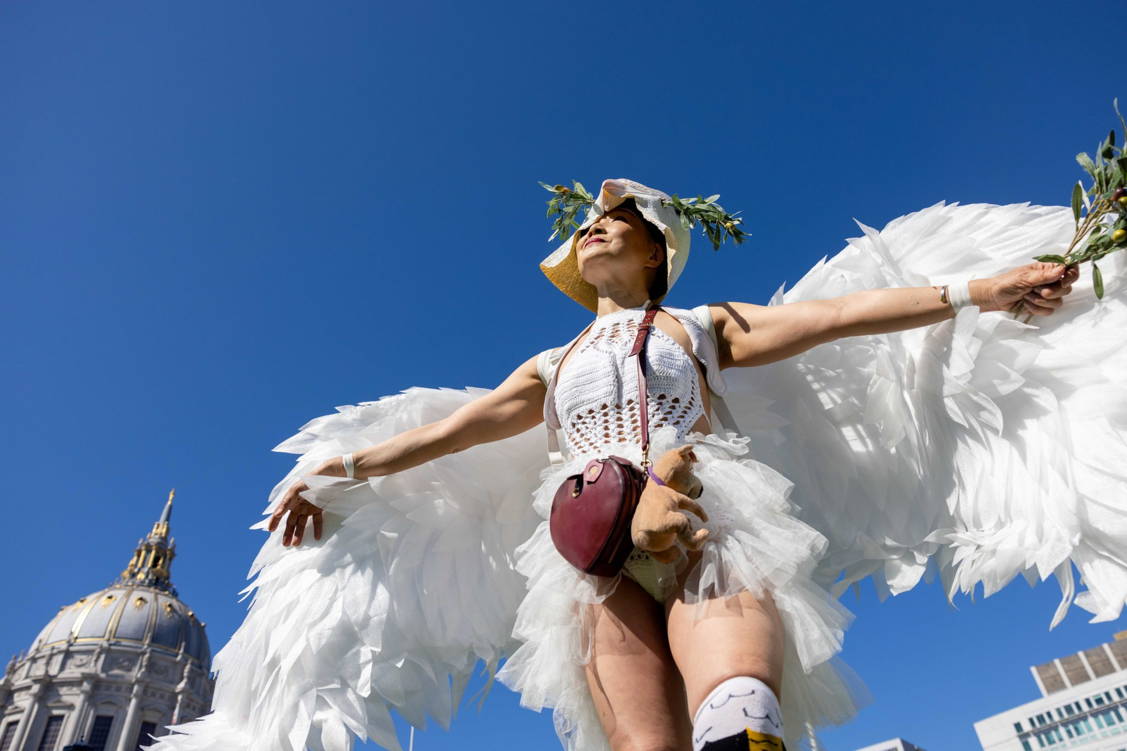 A person wearing large white angel wings, a white outfit, and a hat with greenery stretches their arms holding branches against a clear blue sky.