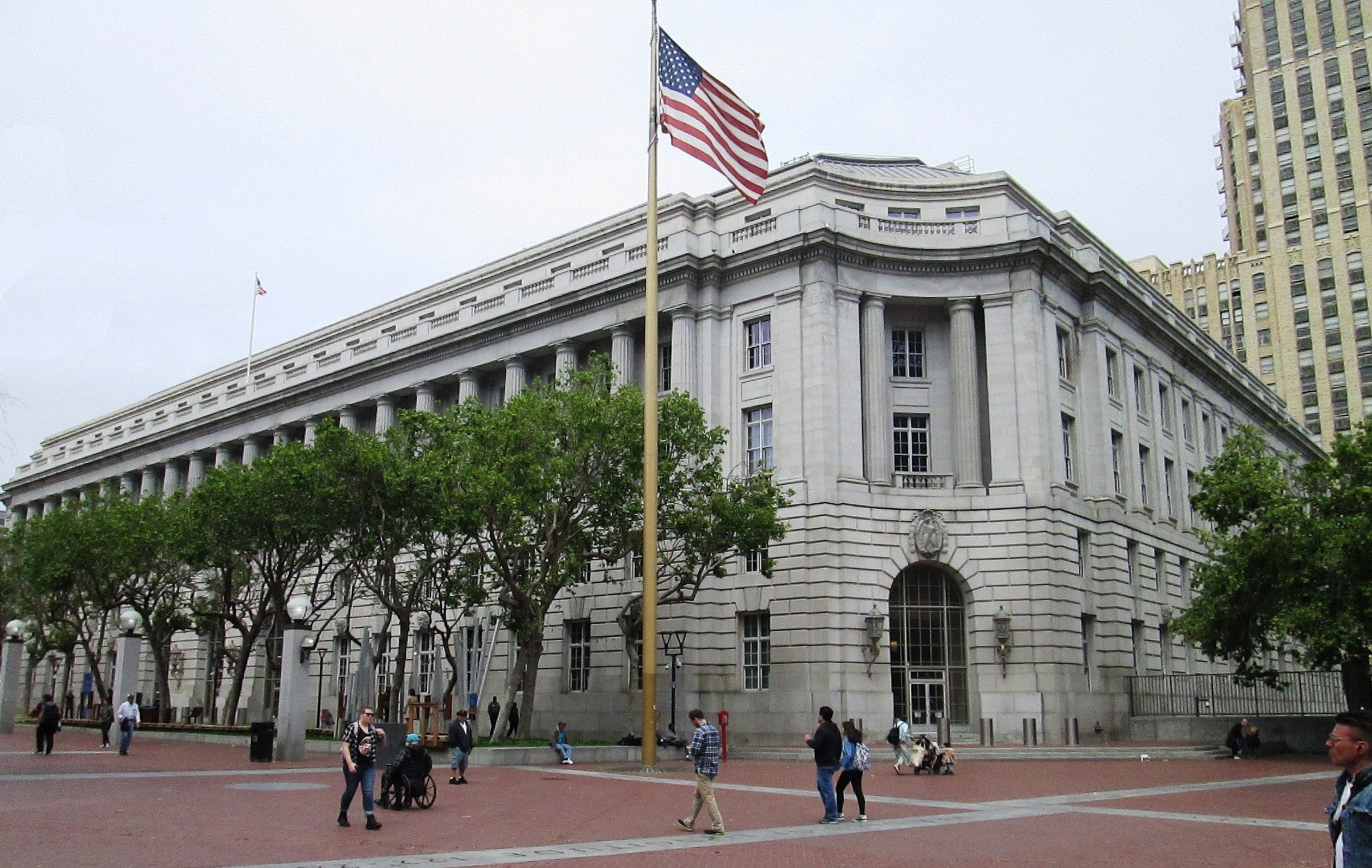 A large neoclassical building with columns is in the background. An American flag flies prominently in front. People walk and gather in the open plaza.