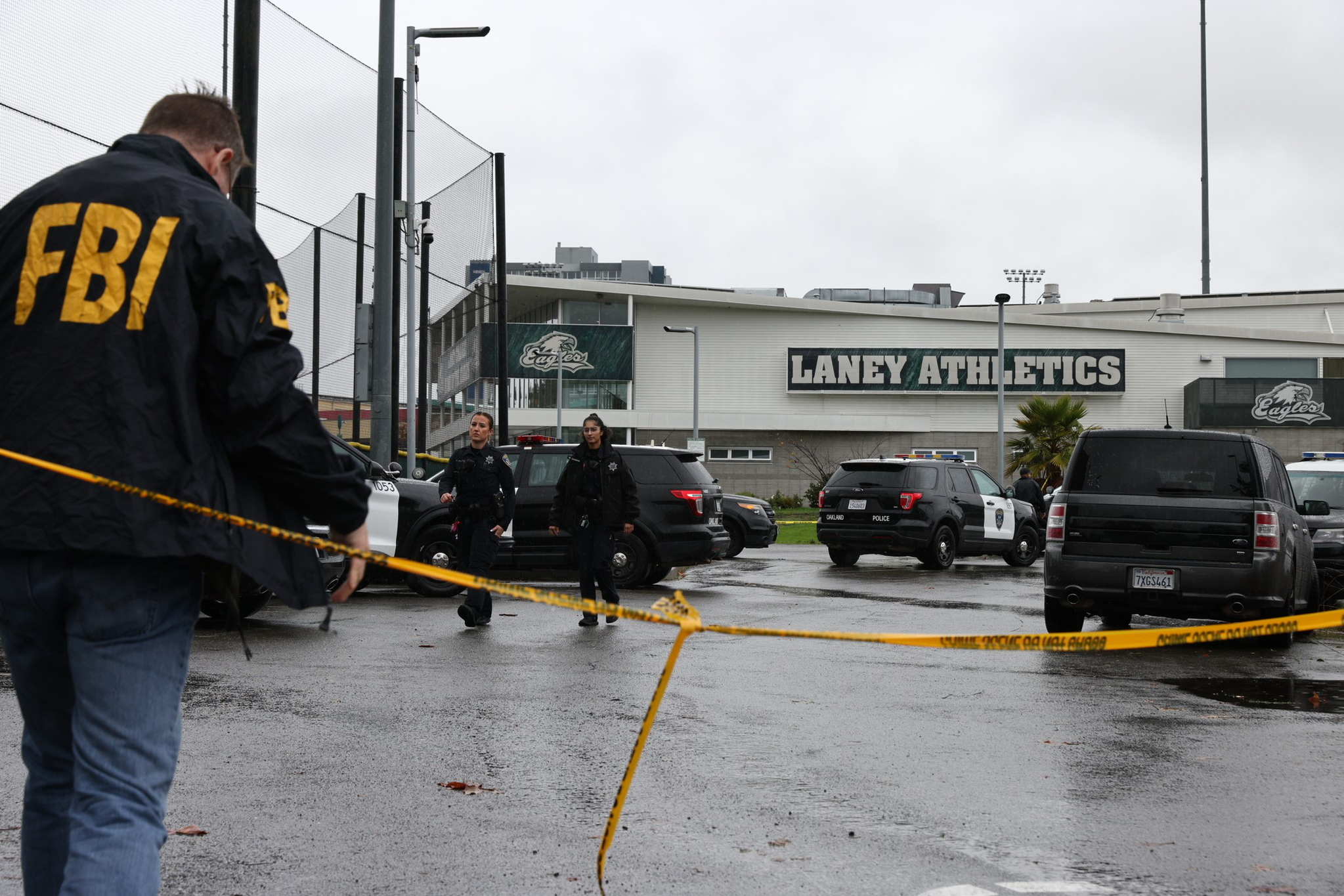 An FBI agent and two police officers secure a wet parking lot outside Laney Athletics building with yellow caution tape and police vehicles present.