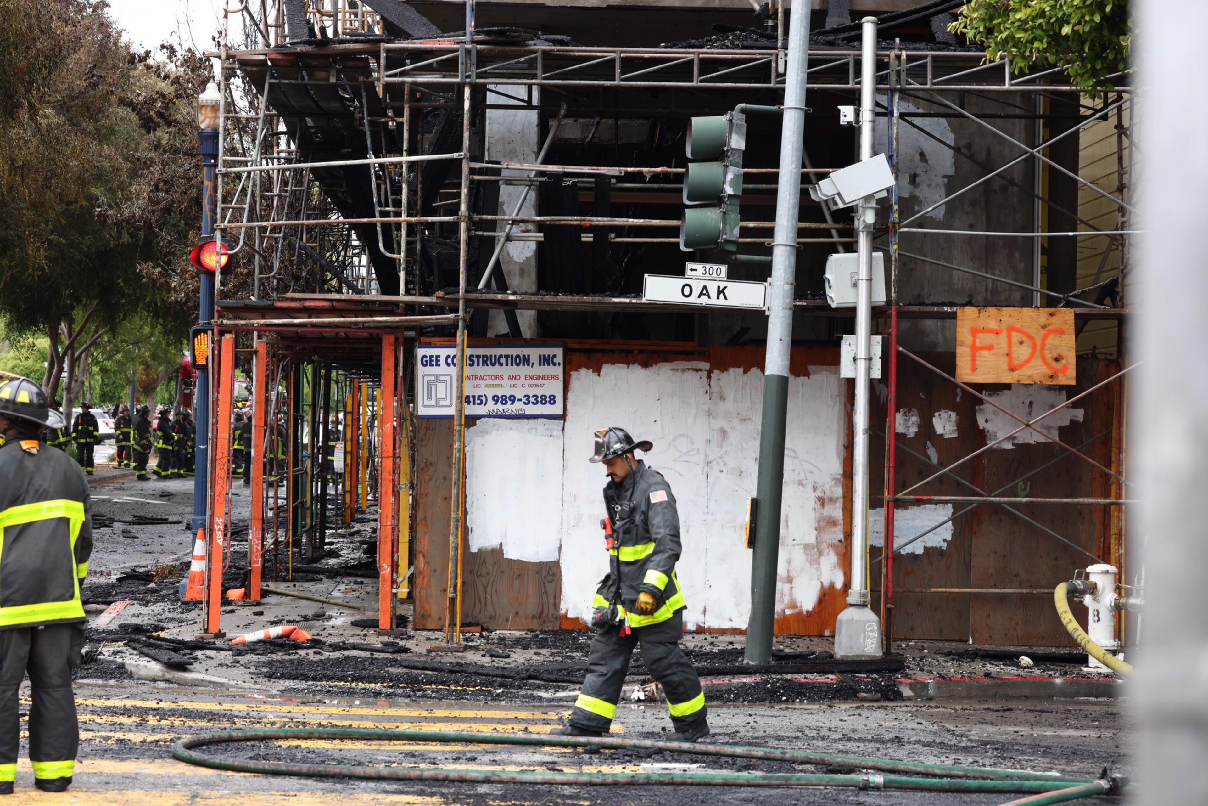 A firefighter walks across a street covered in debris and hoses near a building under construction with scaffolding and fire damage.