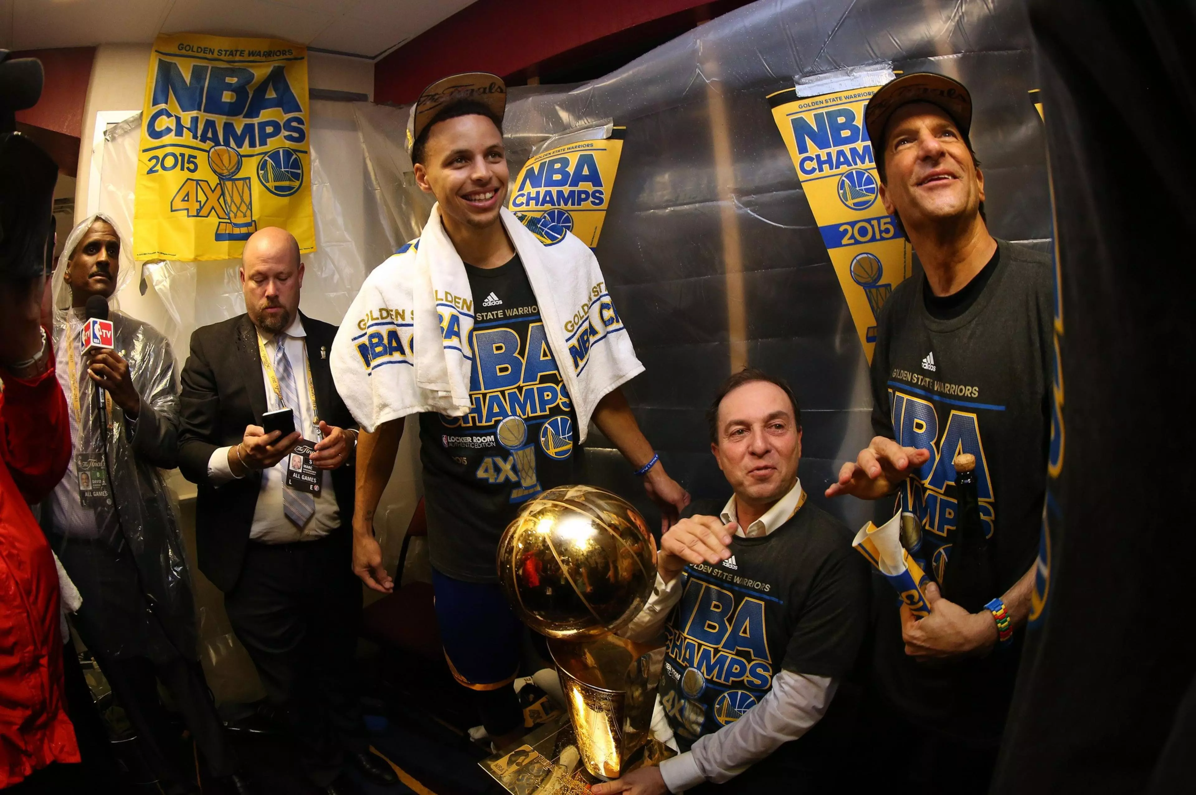 Golden State Warriors players and staff celebrate their 2015 NBA Championship with a trophy, wearing NBA Champs shirts and caps in a locker room.