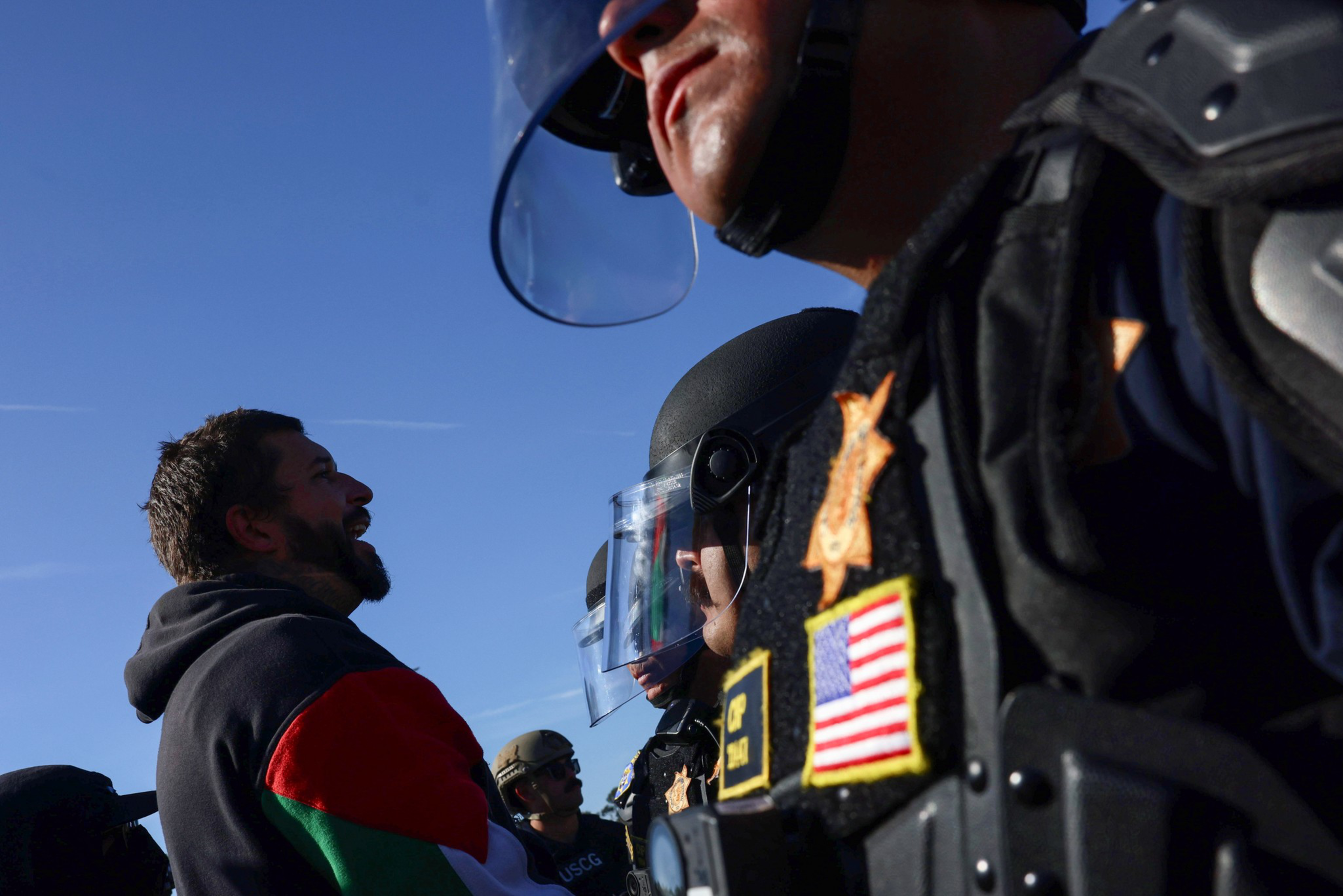 A protester with a beard speaks intensely facing a line of police officers wearing helmets, face shields, and tactical gear against a clear blue sky.