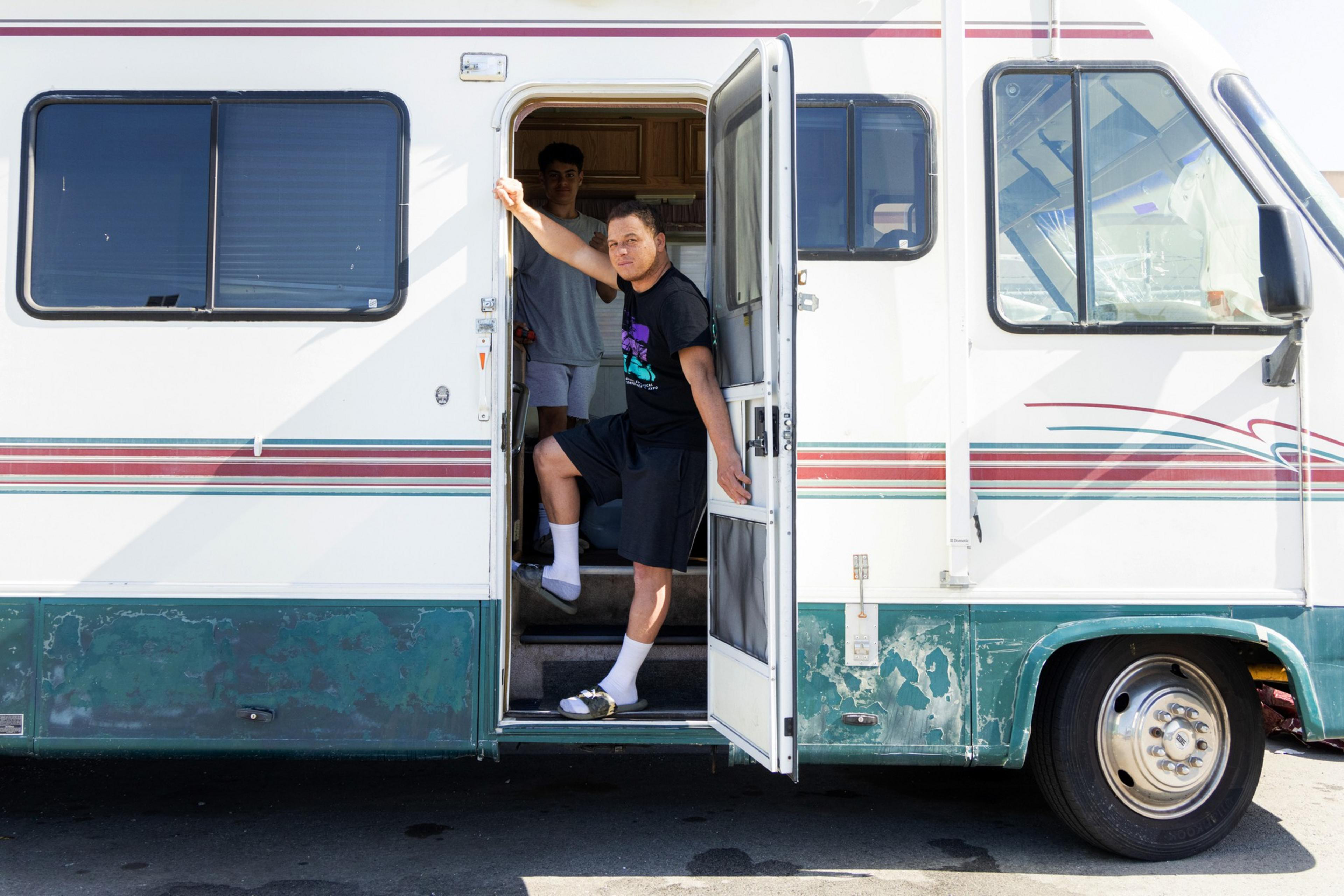 Two people are at the open door of an old RV; one stands outside, leaning on the door frame, while the other is inside, partially visible.