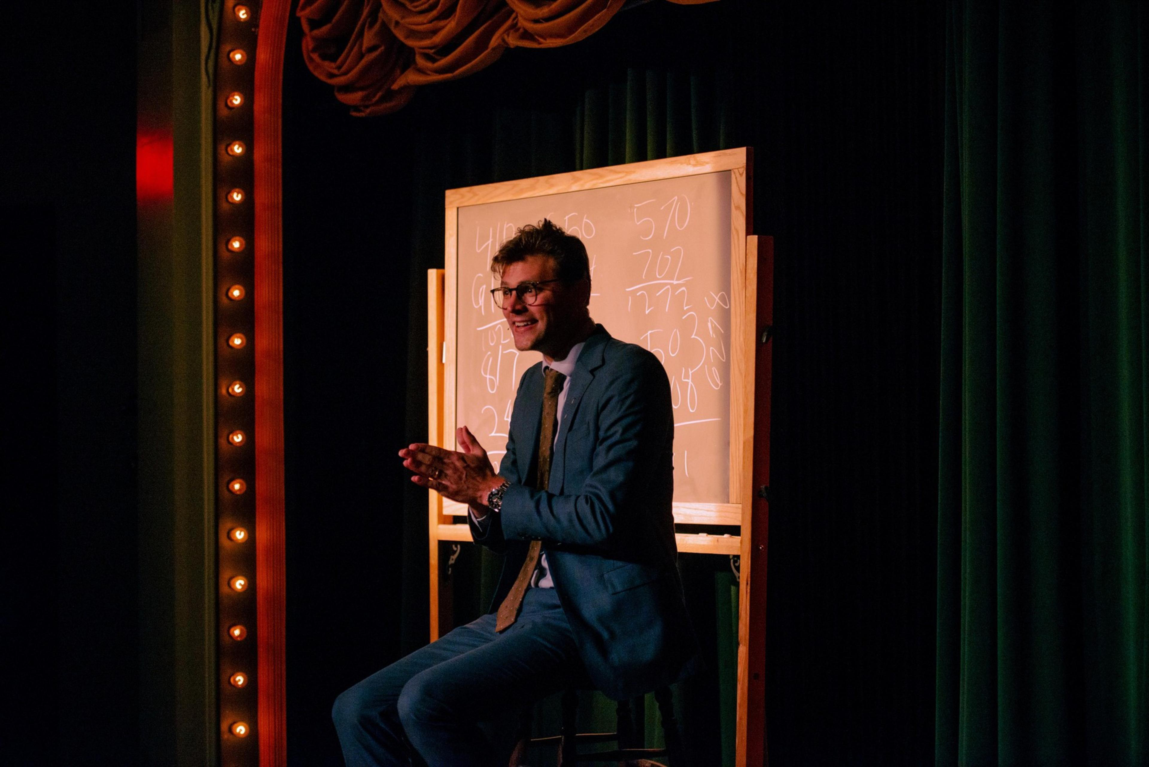 A man in a blue suit and tie sits on a stool, smiling and clapping, with a whiteboard covered in numbers behind him on a dimly lit stage.