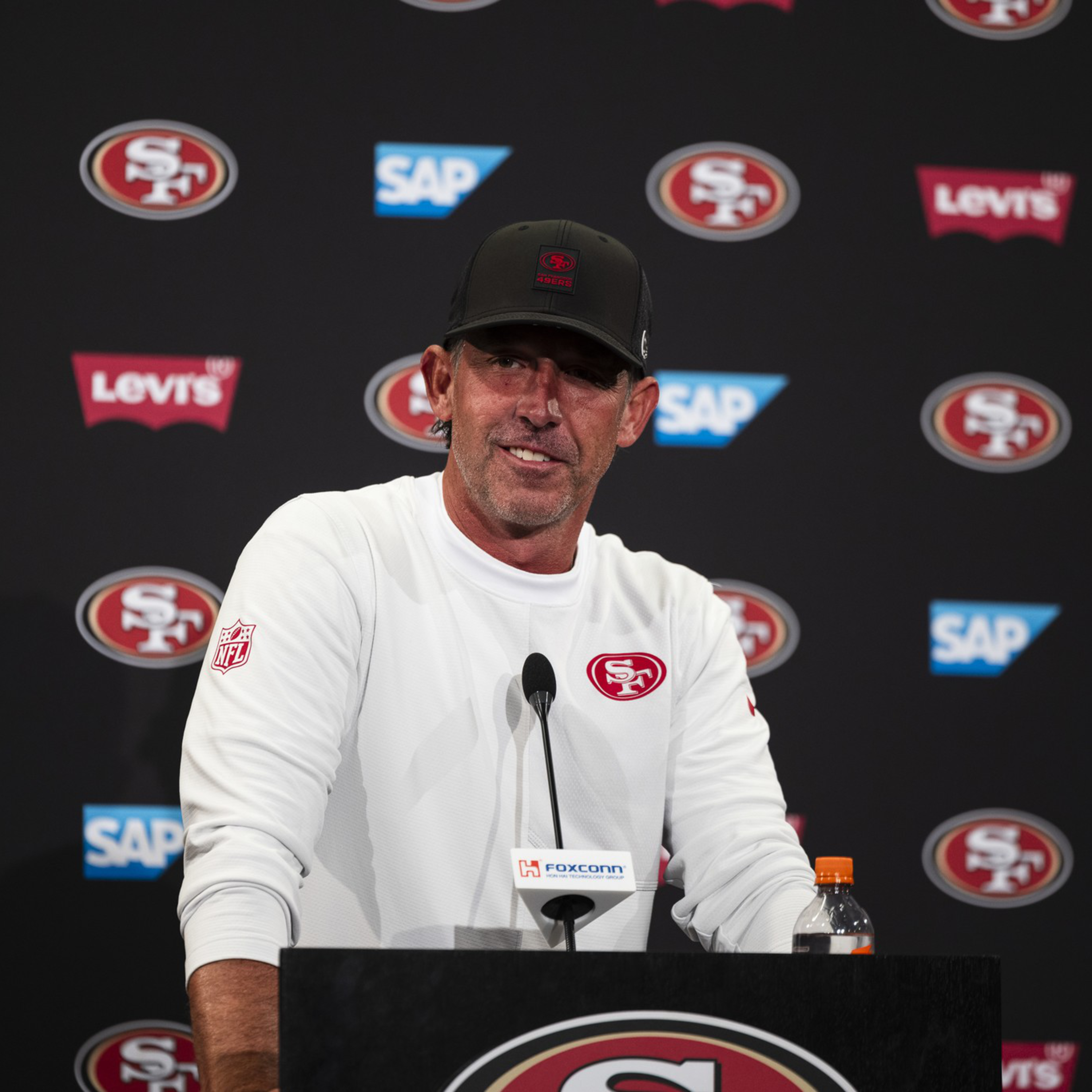 A man wearing a white San Francisco 49ers shirt and black cap speaks at a podium with 49ers, Levi's, and SAP logos in the background.