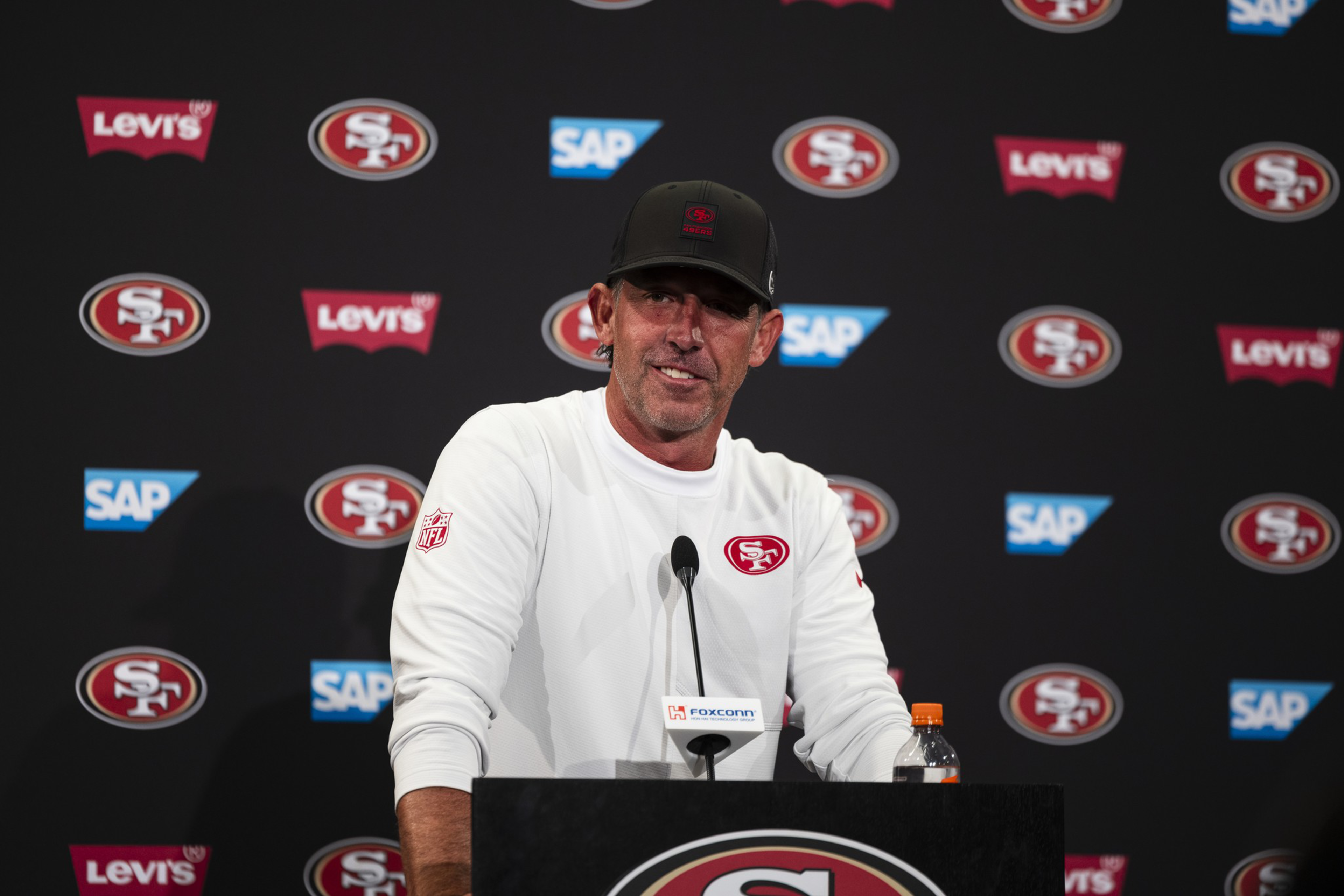 A man wearing a white San Francisco 49ers shirt and black cap speaks at a podium with 49ers, Levi's, and SAP logos in the background.