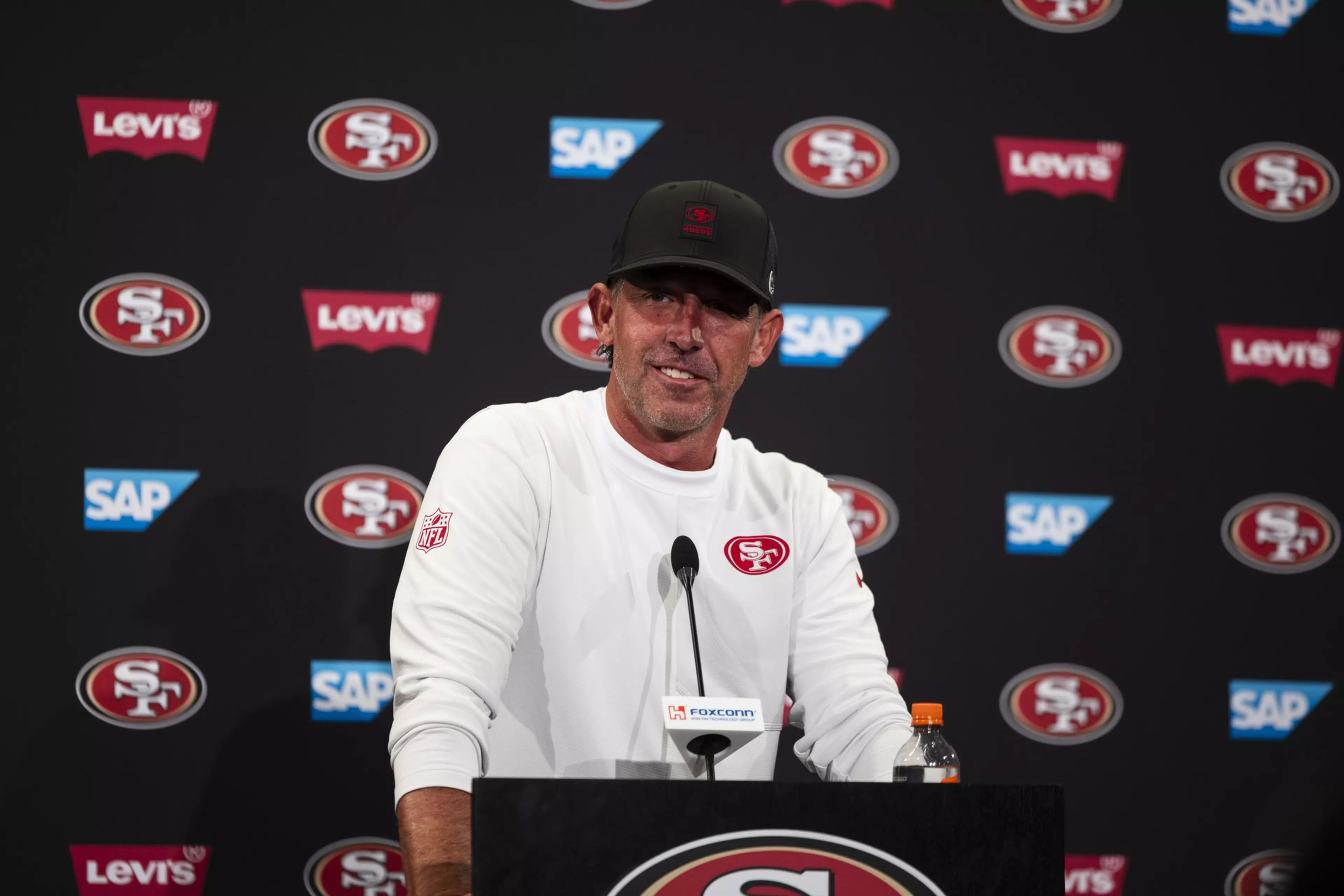 A man wearing a white San Francisco 49ers shirt and black cap speaks at a podium with 49ers, Levi's, and SAP logos in the background.