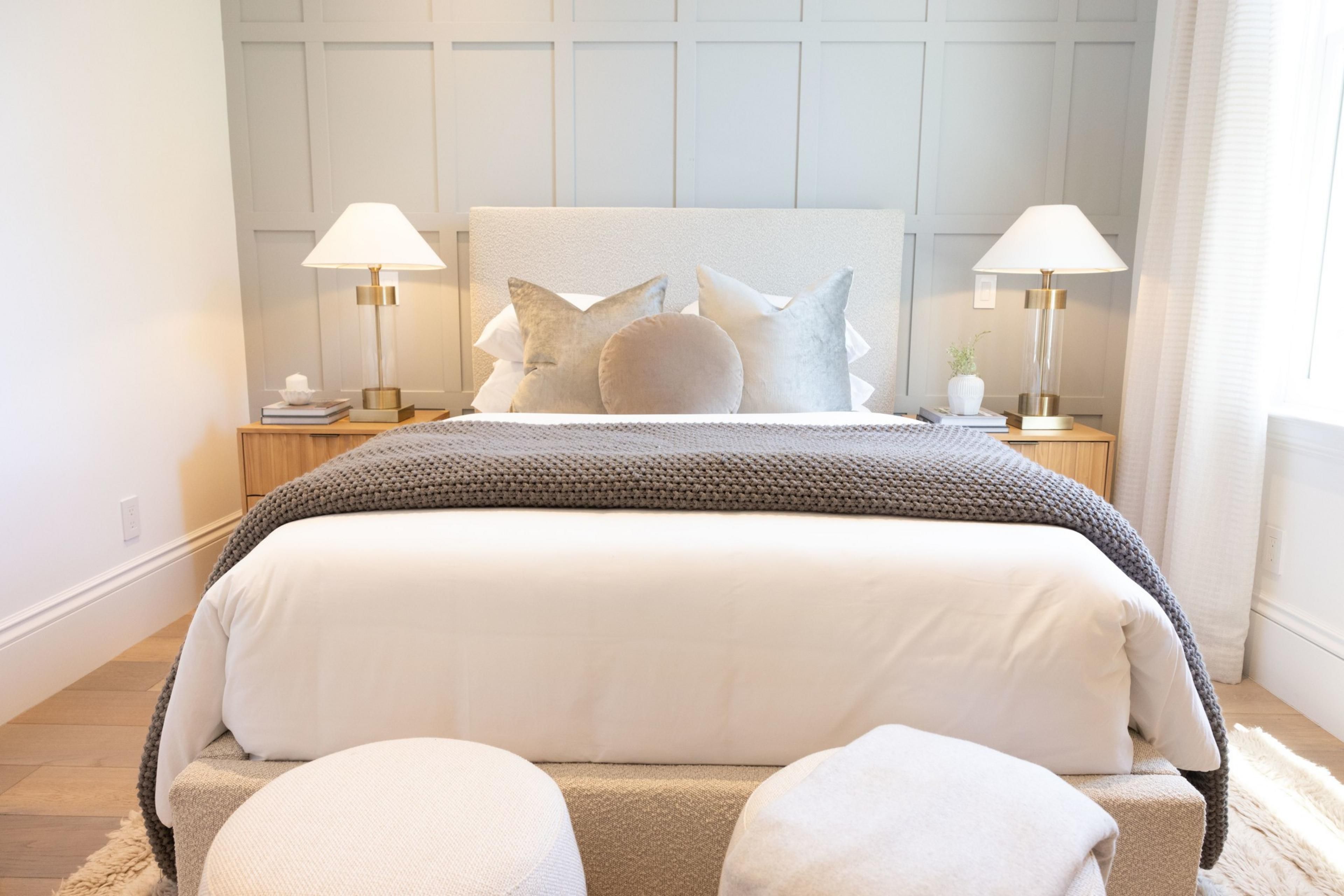 The image shows a neatly made bed with a white headboard, grey pillows, a round throw pillow, and a grey-knit blanket. Two side tables with lamps and decorative items are beside the bed.