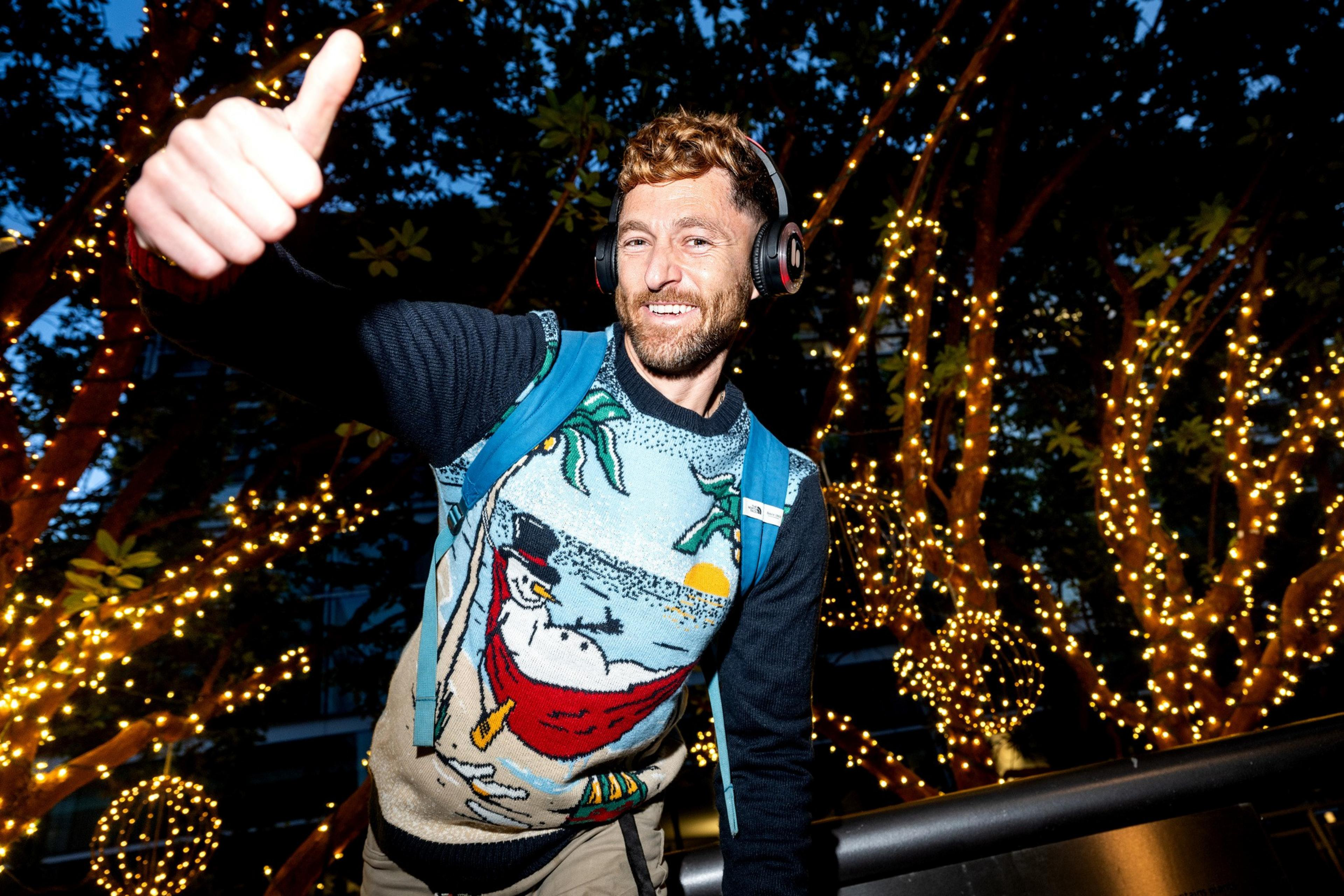 A man with headphones, wearing a festive sweater and backpack, gives a thumbs up in front of trees decorated with twinkling lights.