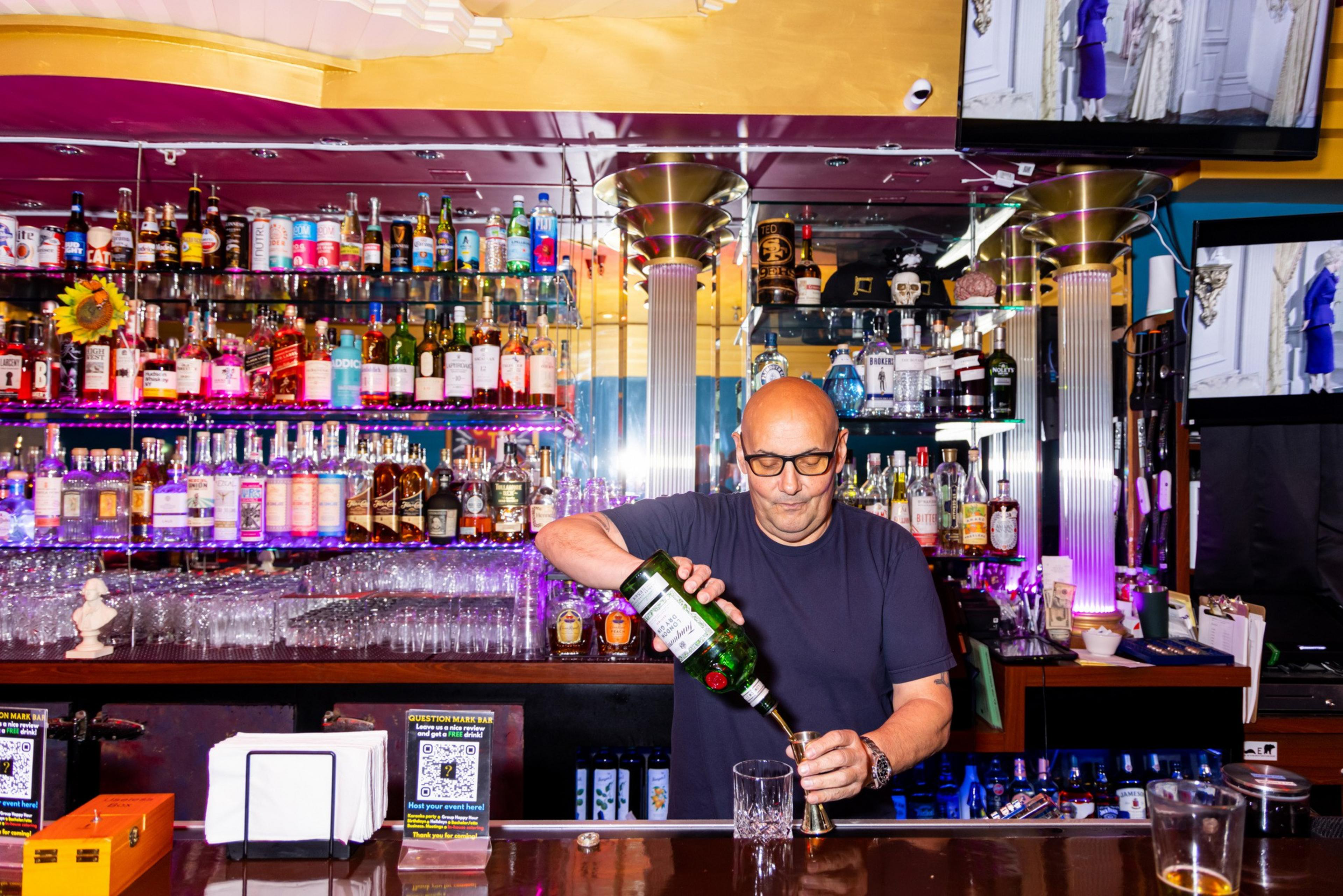 A bartender with glasses pours a green bottle into a jigger at a bar stocked with colorful liquor bottles. Two TV screens are in the background.