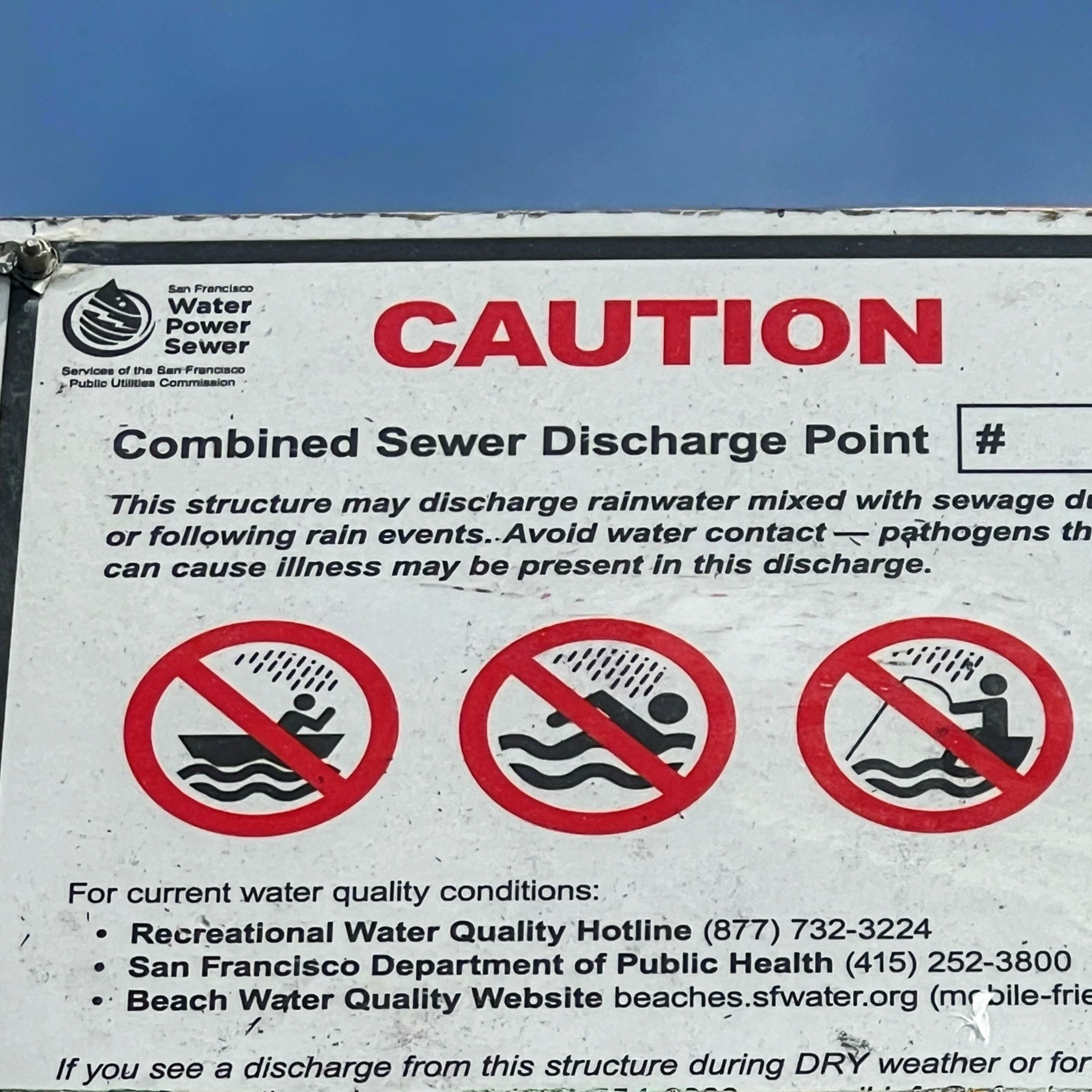 A warning sign indicating a combined sewer discharge point, cautioning against water contact due to possible rainwater and sewage mixing, with contact details for water quality conditions.