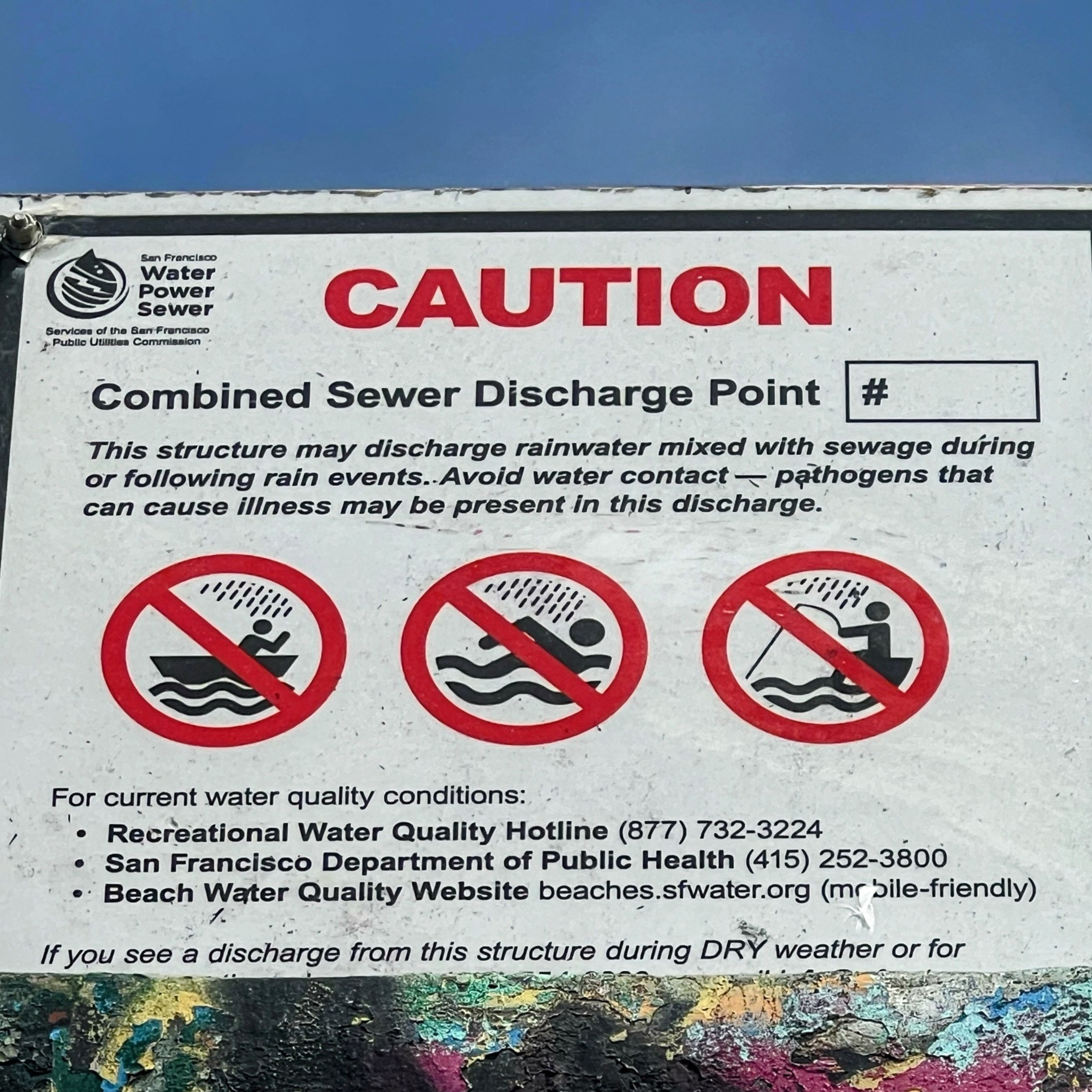 A warning sign indicating a combined sewer discharge point, cautioning against water contact due to possible rainwater and sewage mixing, with contact details for water quality conditions.