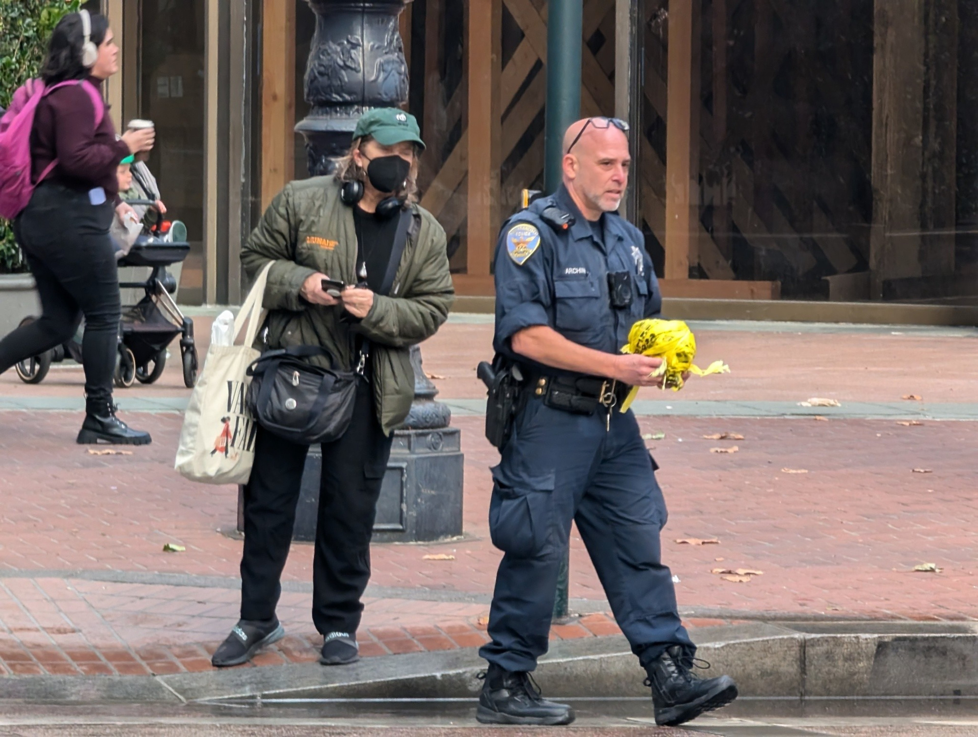 A street scene shows a woman standing at an intersection on a brick sidewalk and wearing a mask and baseball cap next to a police officer holding a rolled-up ball of yellow caution tape that had been cordoning off an investigation area.