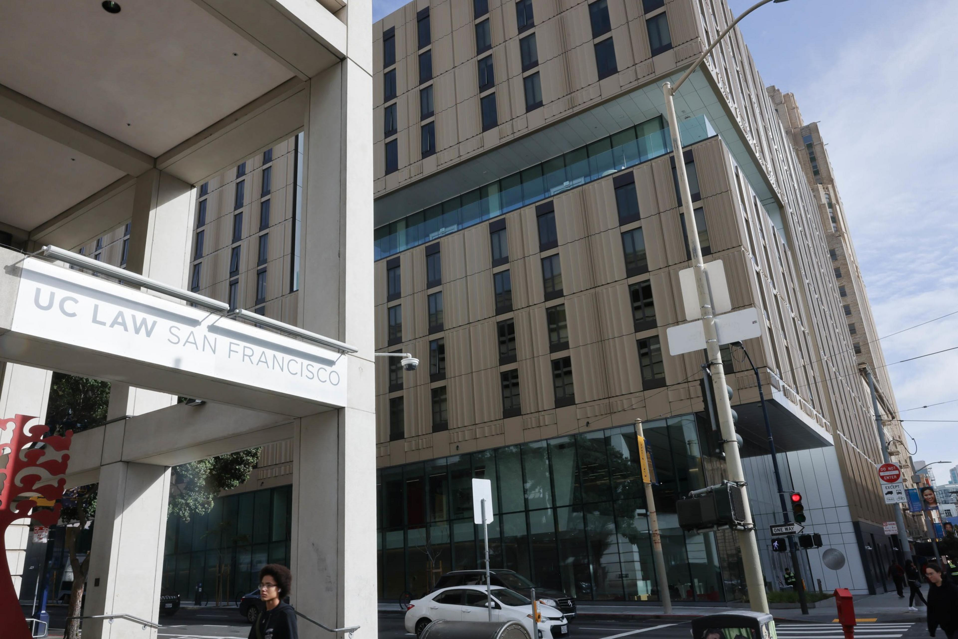 A city street corner with a sign &quot;UC LAW SAN FRANCISCO&quot; and some modern buildings in the background.