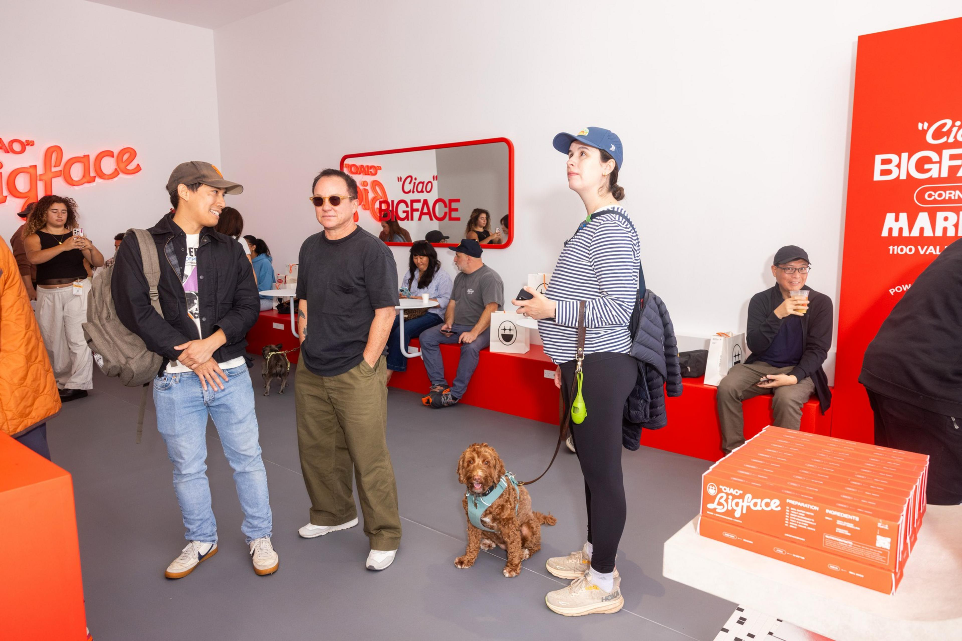The image shows a lively cafe with people chatting and a dog sitting by a woman in a cap. Bright red &quot;Ciao Bigface&quot; signs and decor are prominent.