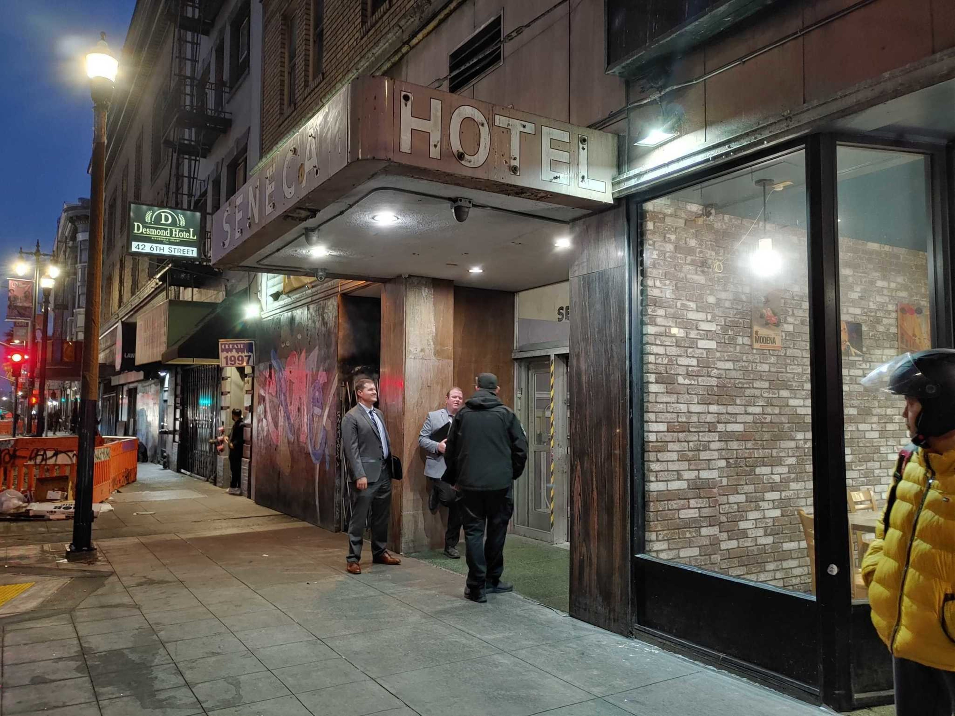 Three men stand outside a dimly lit Seneca Hotel entrance on a city street at dusk, with one person in a yellow jacket and helmet nearby.