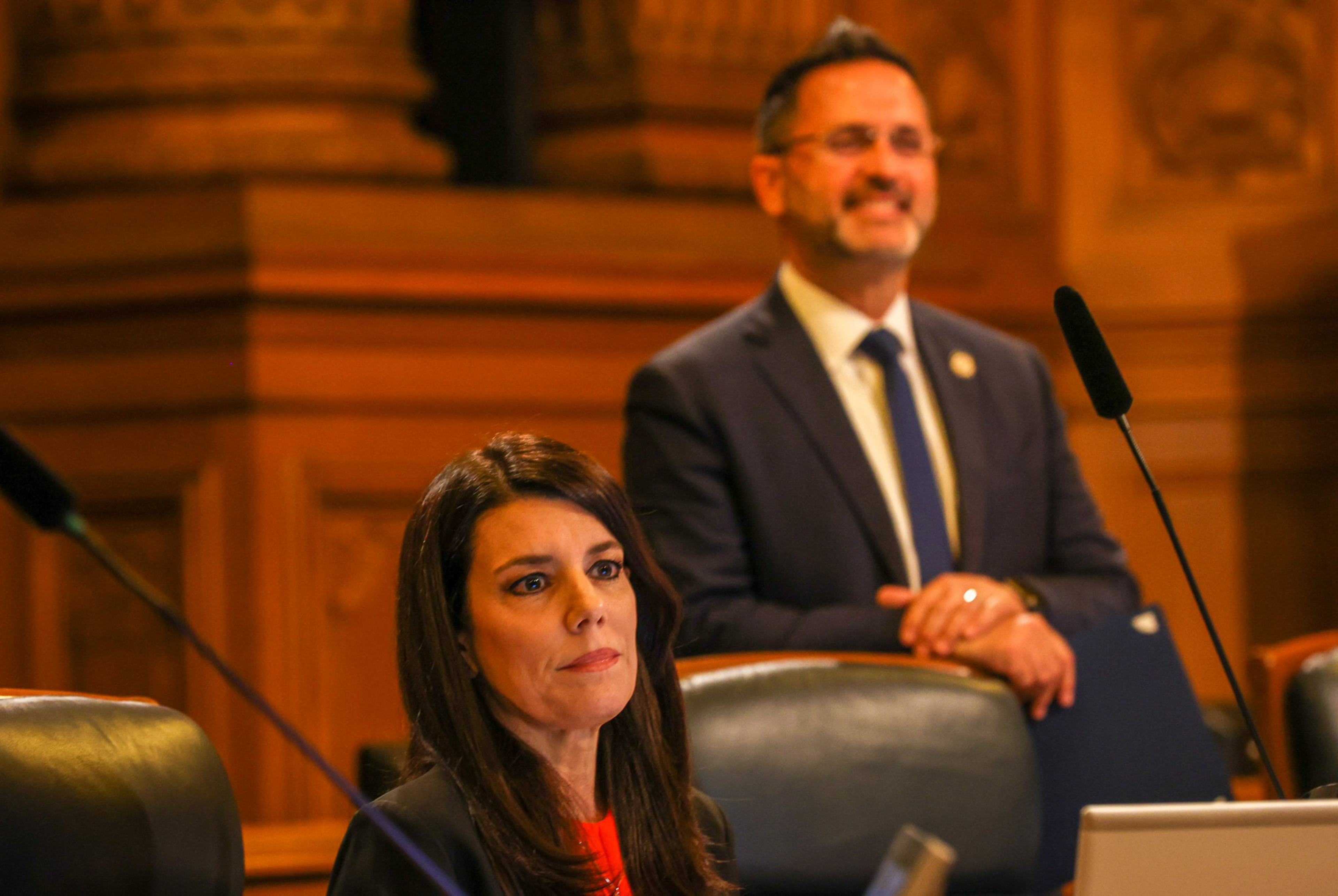 A woman with long dark hair sits attentively at a desk, while a man in a suit and tie stands behind her, smiling with hands clasped.