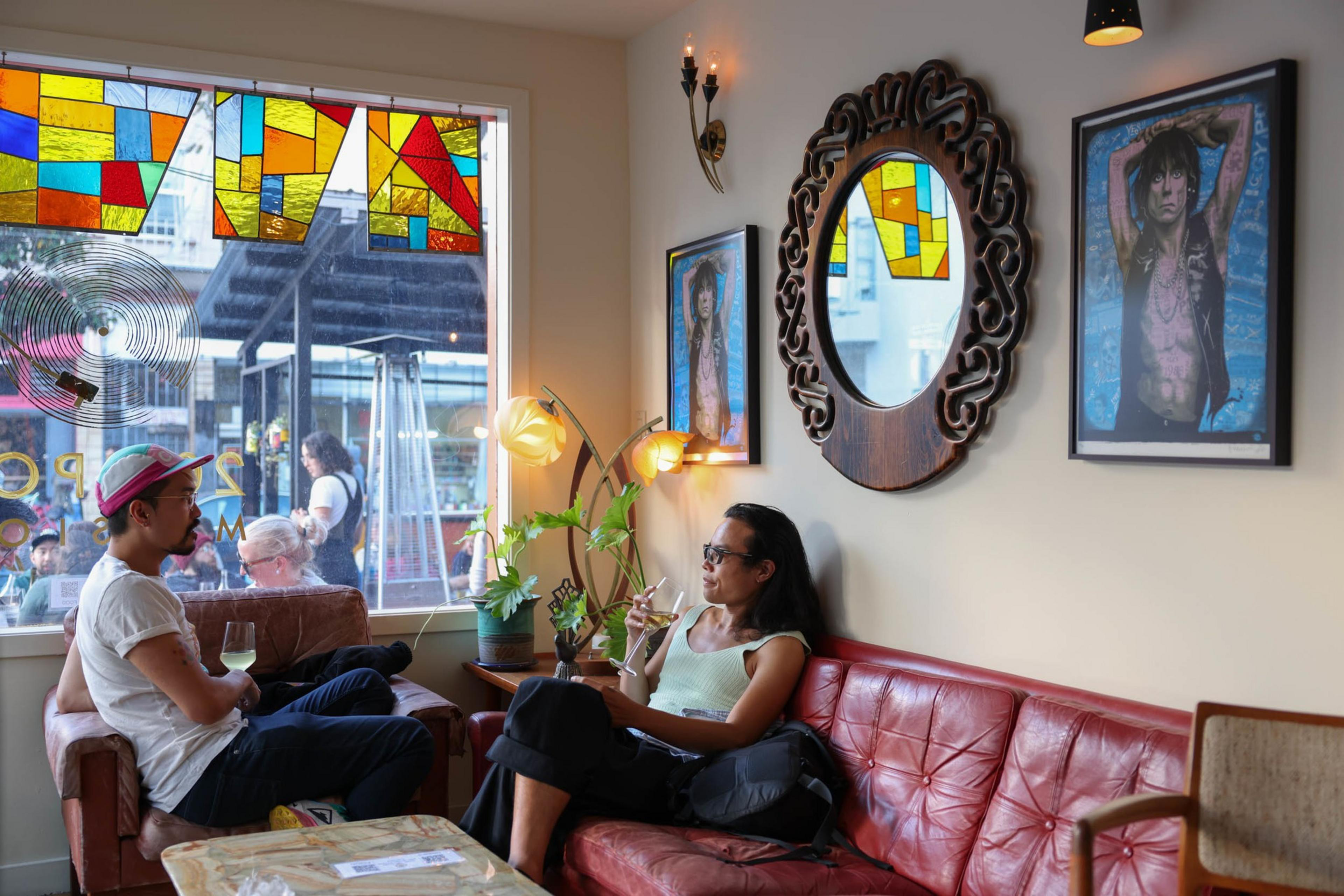 Two people sit in a cozy living room with a stained glass window, a red leather couch, a wooden-framed mirror, and two framed posters on the wall.
