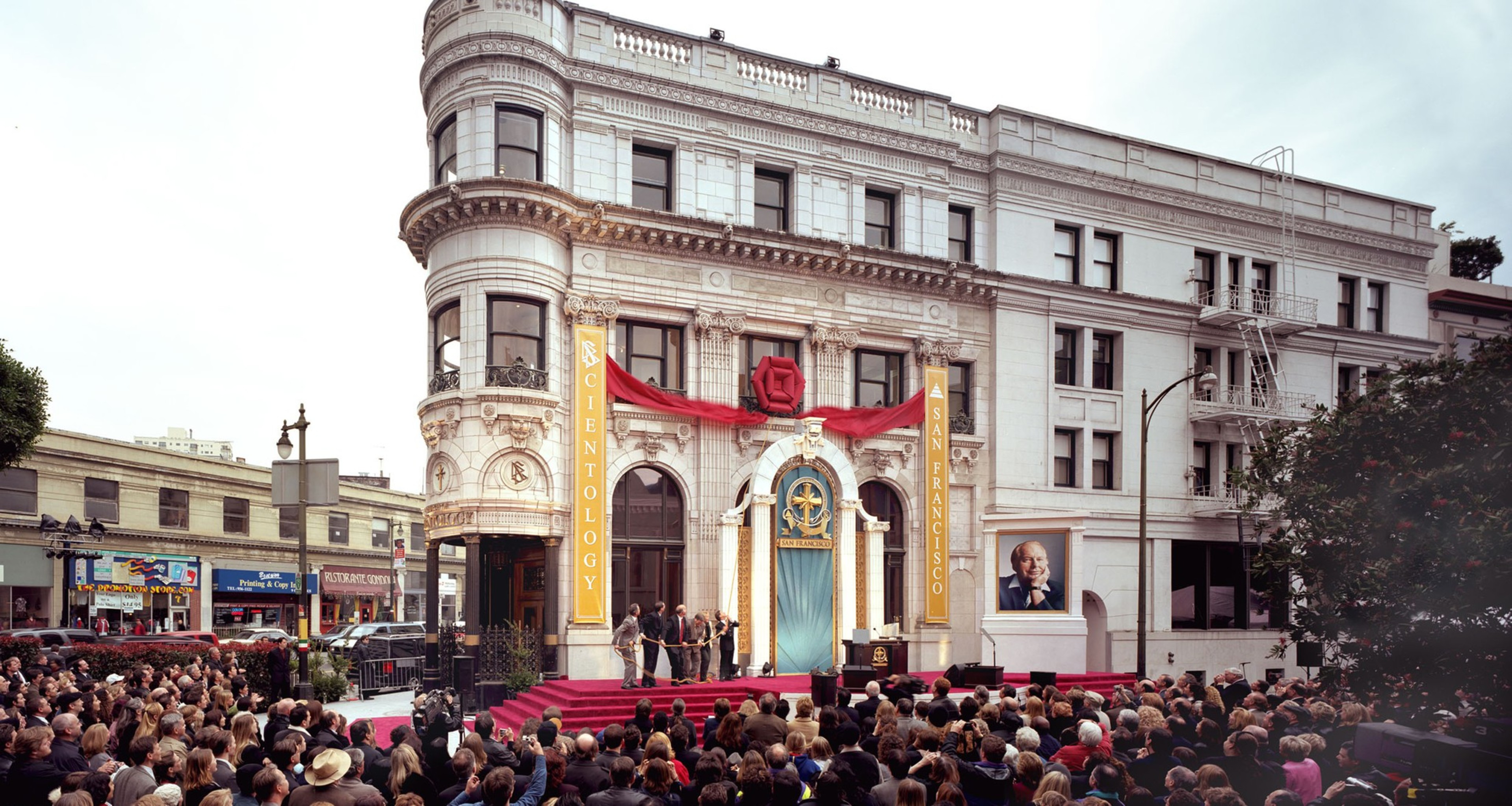 A crowd gathers in front of a grand white building adorned with banners reading &quot;Scientology San Francisco.&quot; A ceremony with several speakers is taking place.