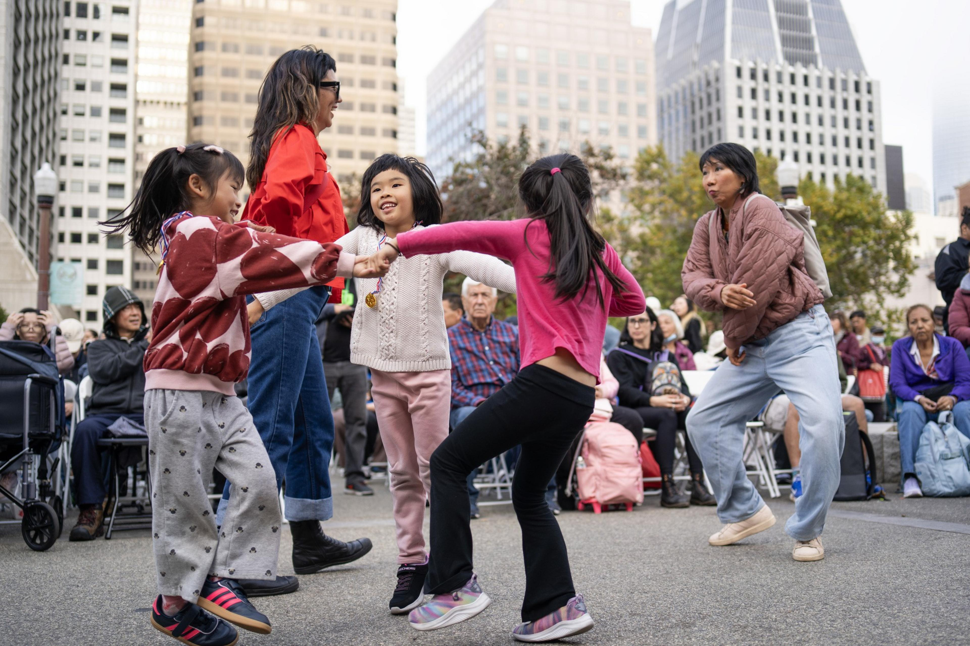 Three children and two adults are holding hands and dancing in a circle outdoors, with a seated crowd and tall buildings in the background.
