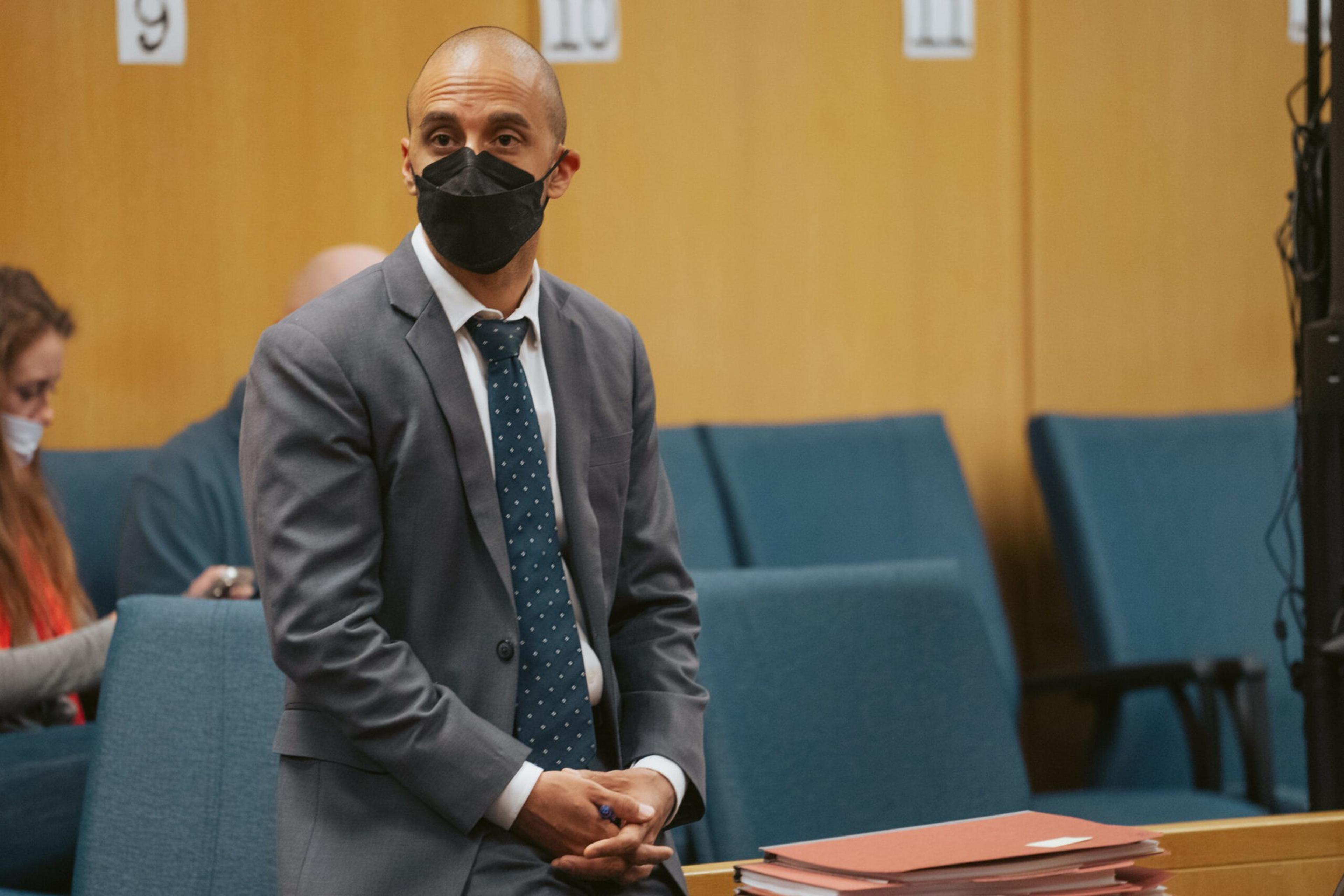 A man in a gray suit and blue polka-dot tie wearing a black mask stands near a stack of documents in a courtroom with blue seats behind him.