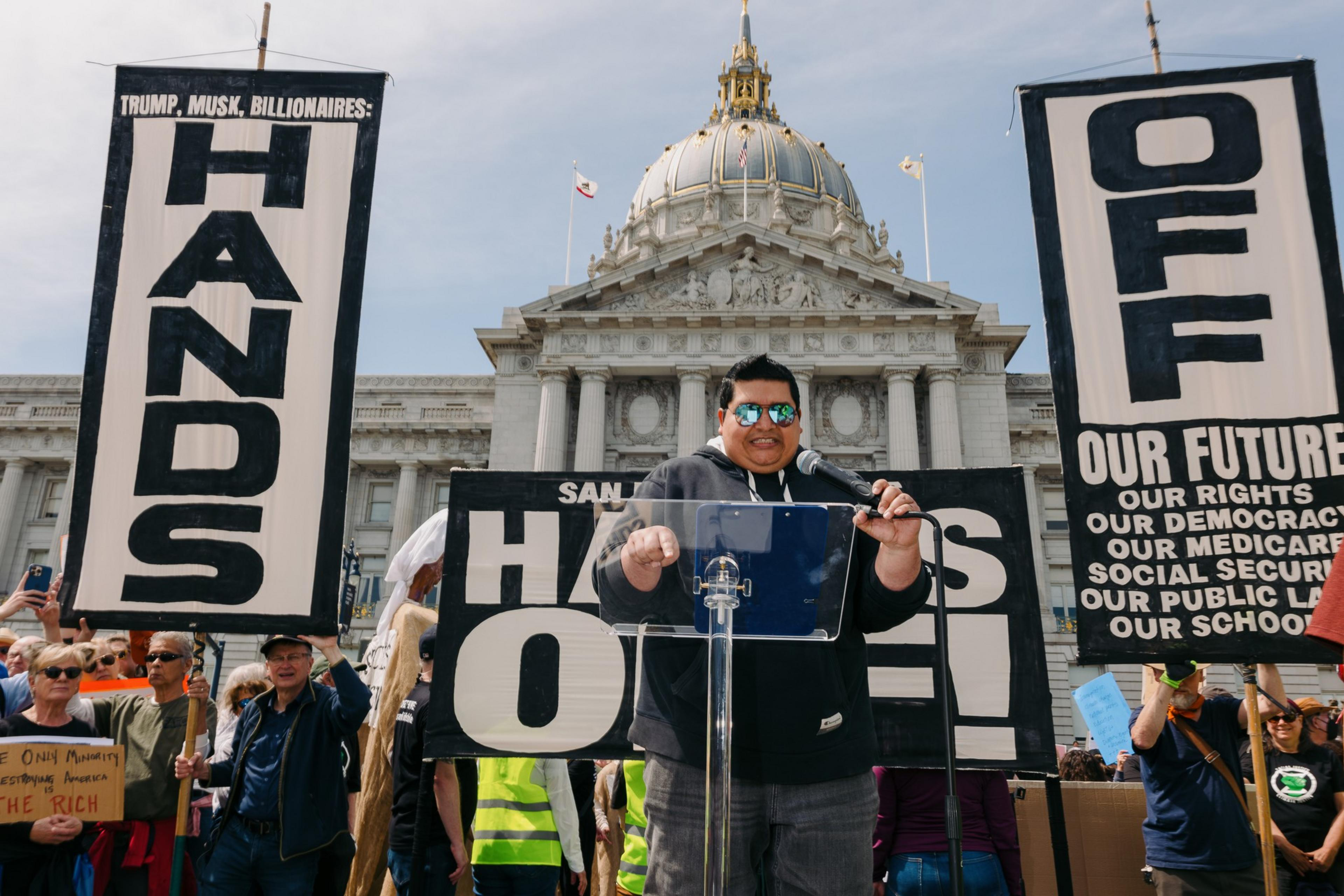 A person speaks at a podium during a protest outside a large, ornate building. The crowd holds signs reading &quot;HANDS OFF&quot; with messages about protecting rights.