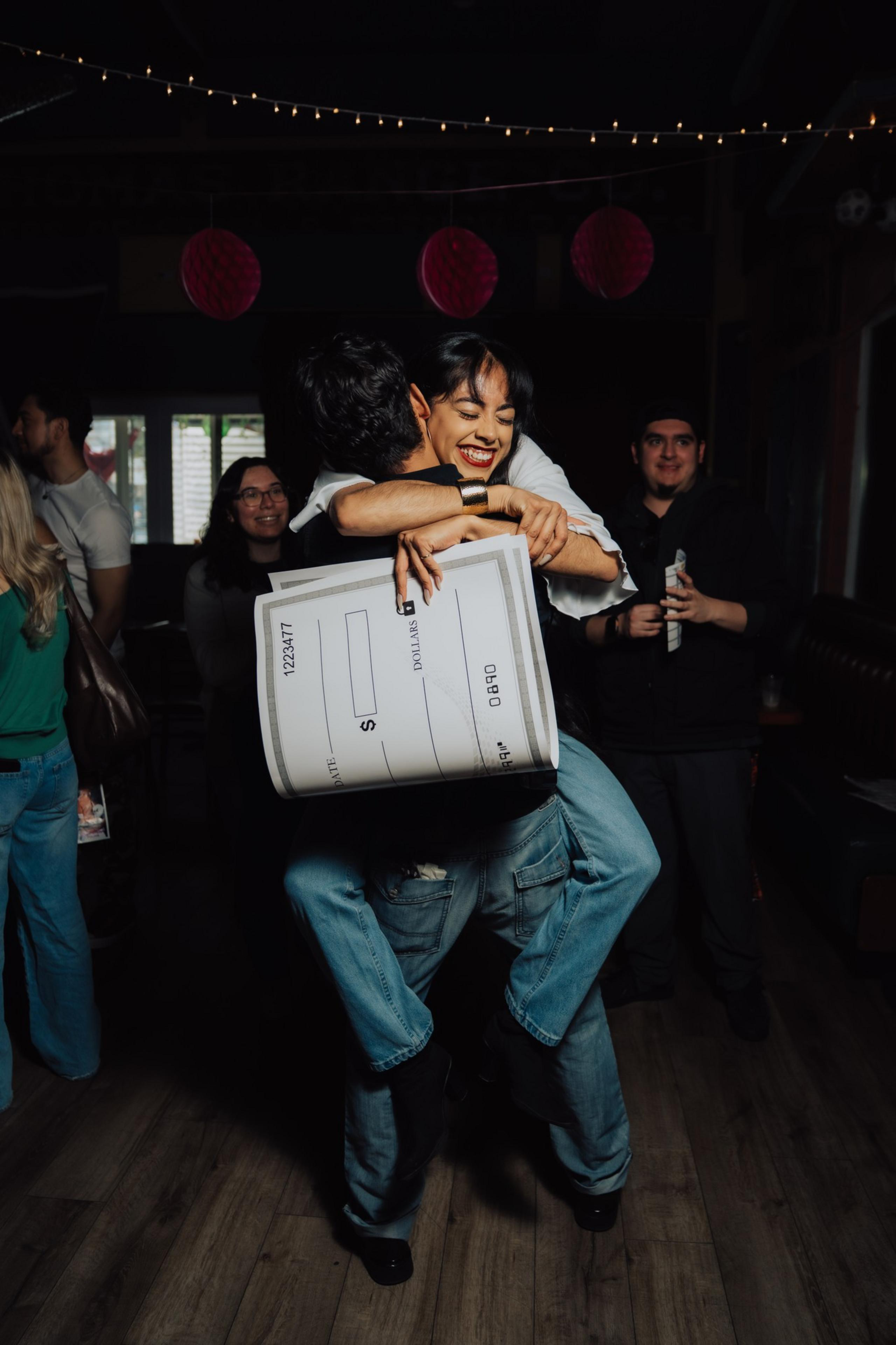 A joyful woman hugs a man tightly while holding a large, blank check. They're surrounded by happy onlookers in a warmly lit room with string lights above.