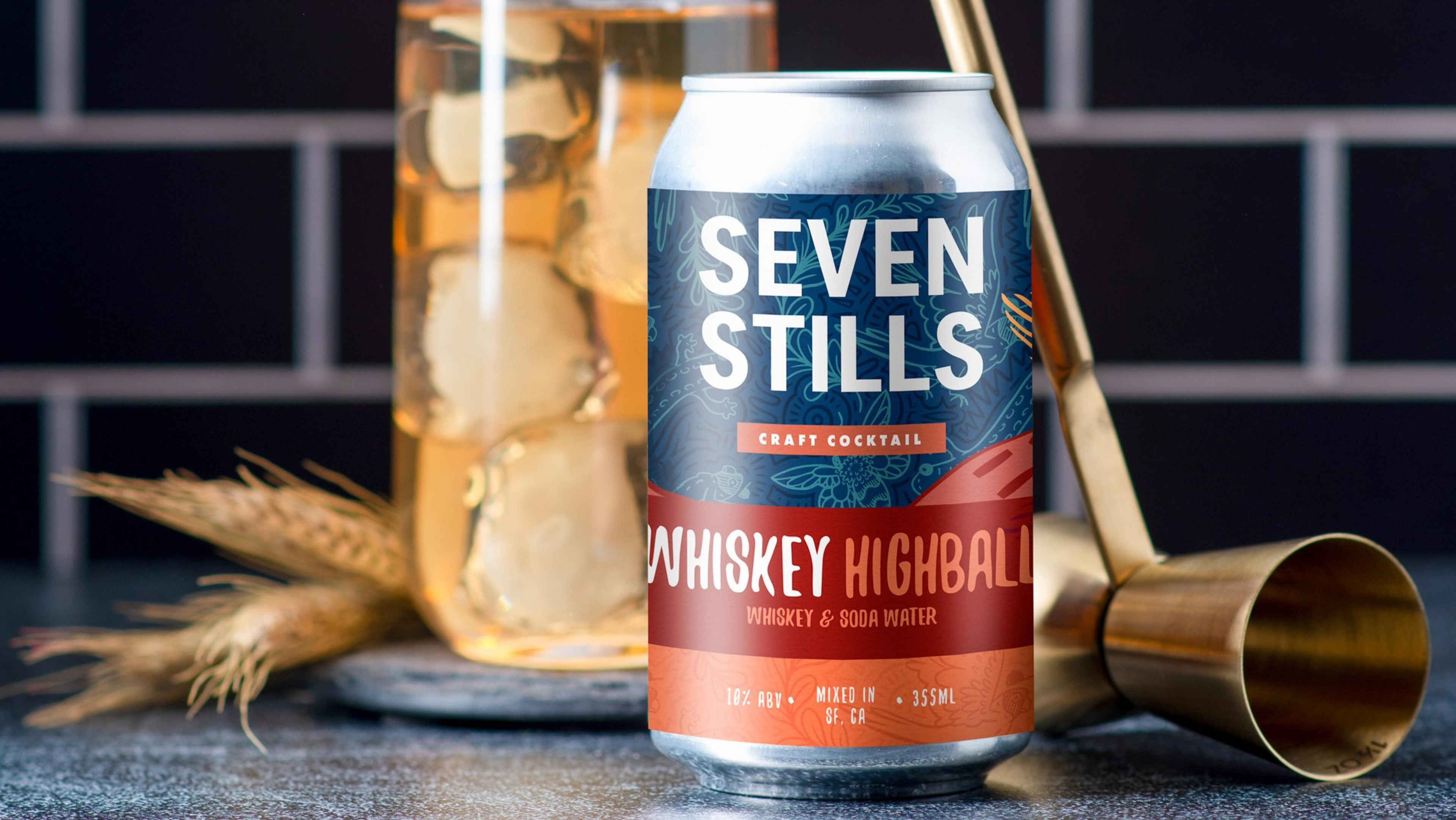 A can of Seven Stills Whiskey Highball craft cocktail sits on a counter with a gold jigger, wheat stalks, and a glass filled with ice and whiskey soda behind it.