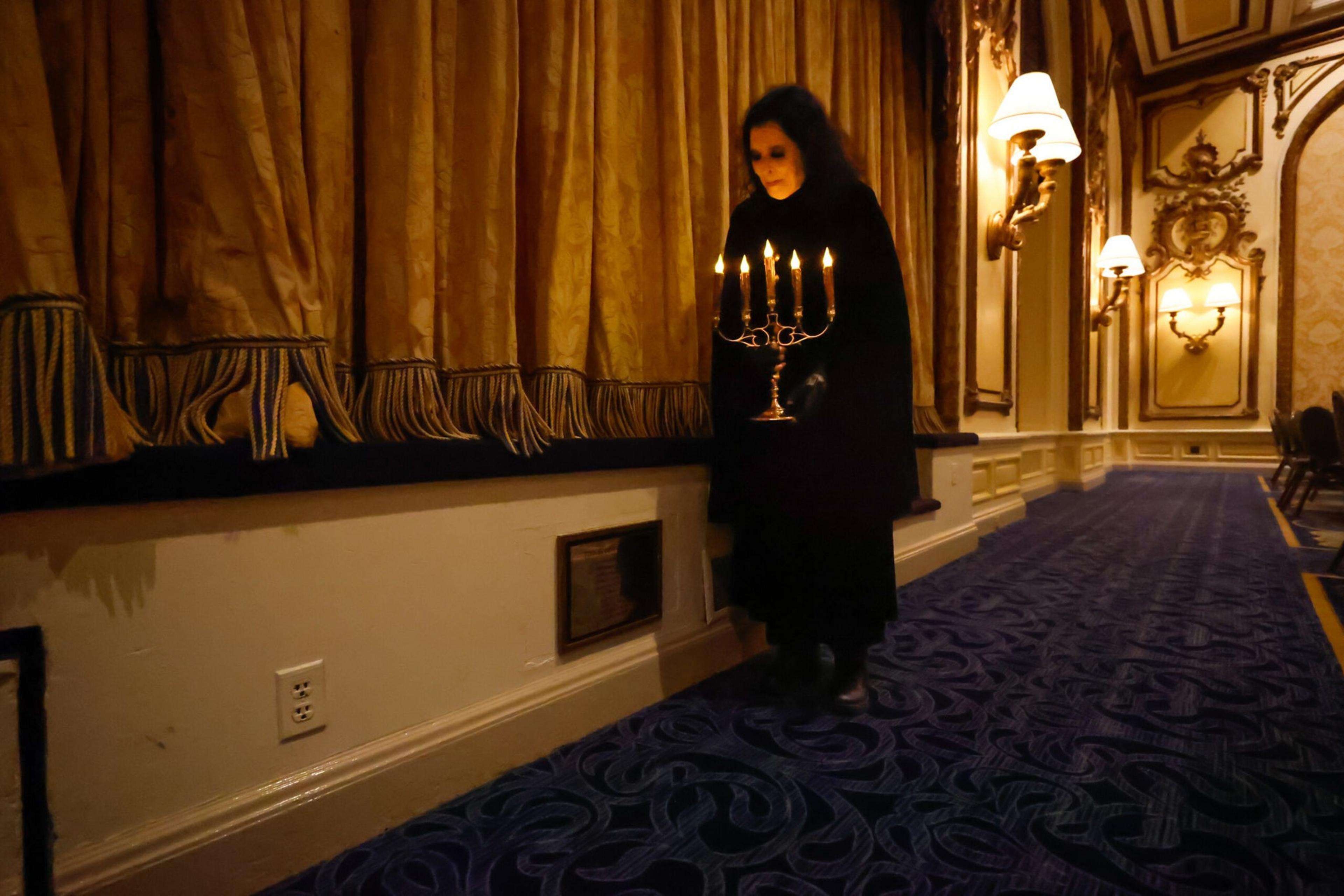 A person dressed in black holds a lit candelabrum walking along a dimly lit, ornate hallway with blue carpet and golden curtains.