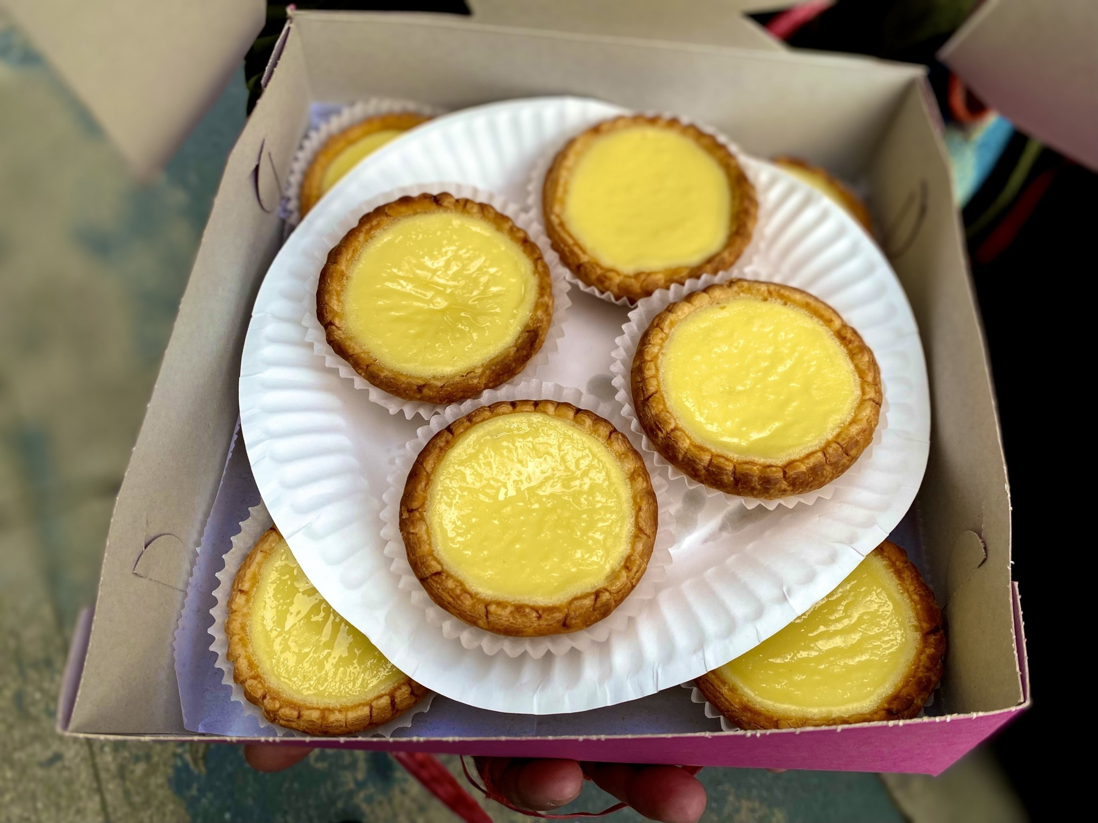 Seven golden-brown egg tarts with smooth yellow custard filling rest on white paper liners inside a cardboard box.