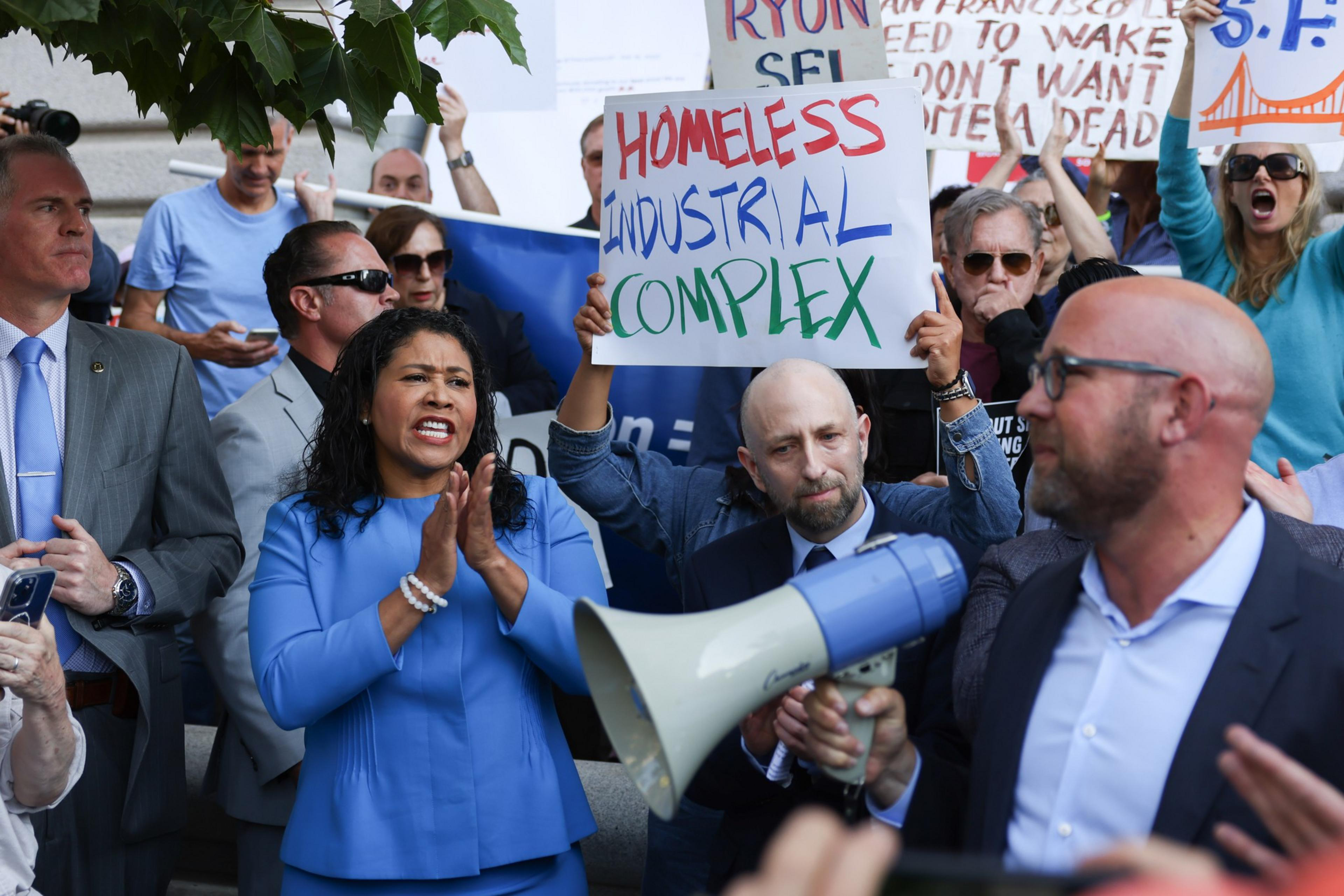 A group of people, including a woman in a blue suit speaking and clapping, gather with protest signs about homelessness. One sign reads &quot;Homeless Industrial Complex.&quot;