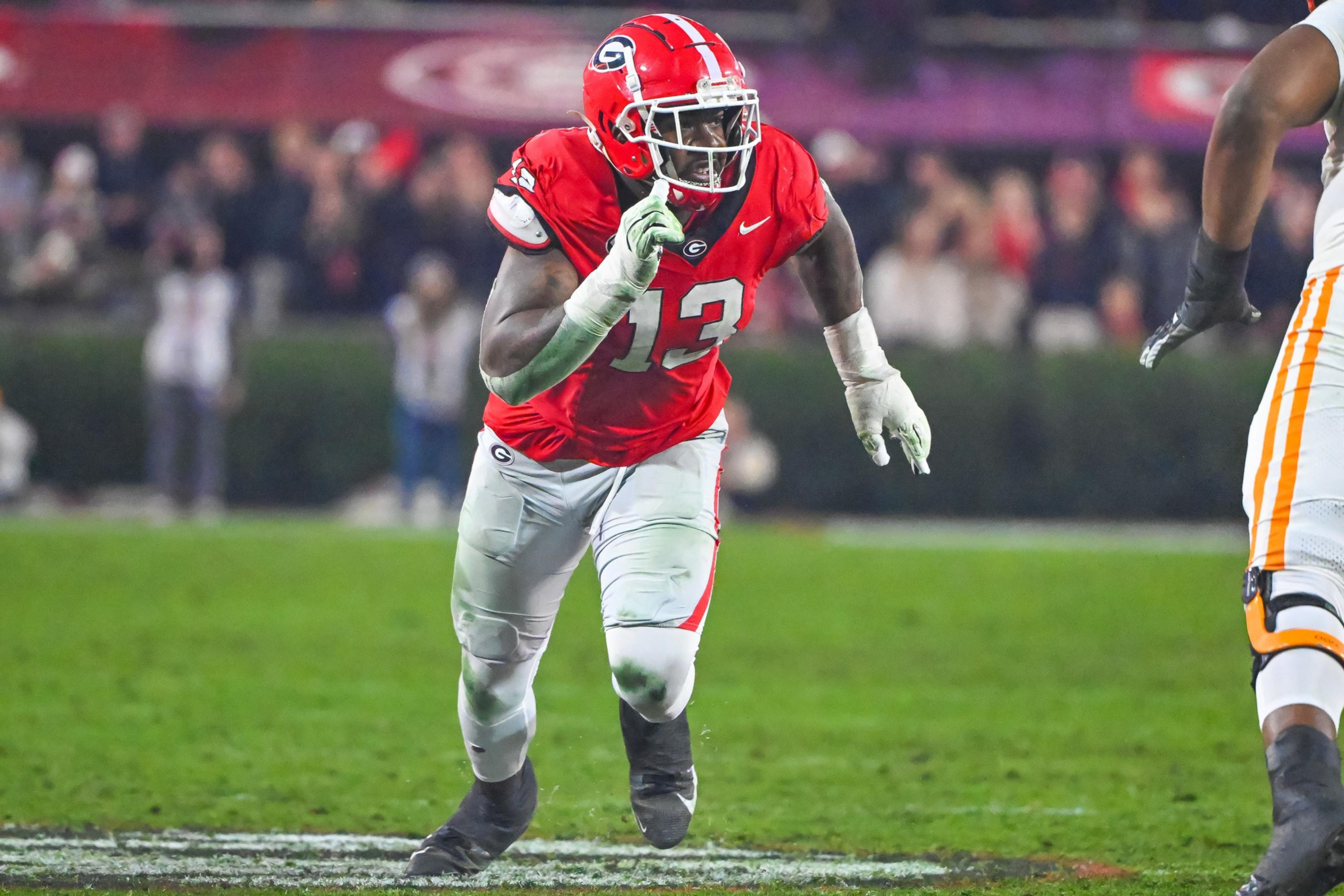 A football player in a red jersey and white pants is mid-action on a field. He wears a helmet with a logo and is surrounded by blurred spectators.