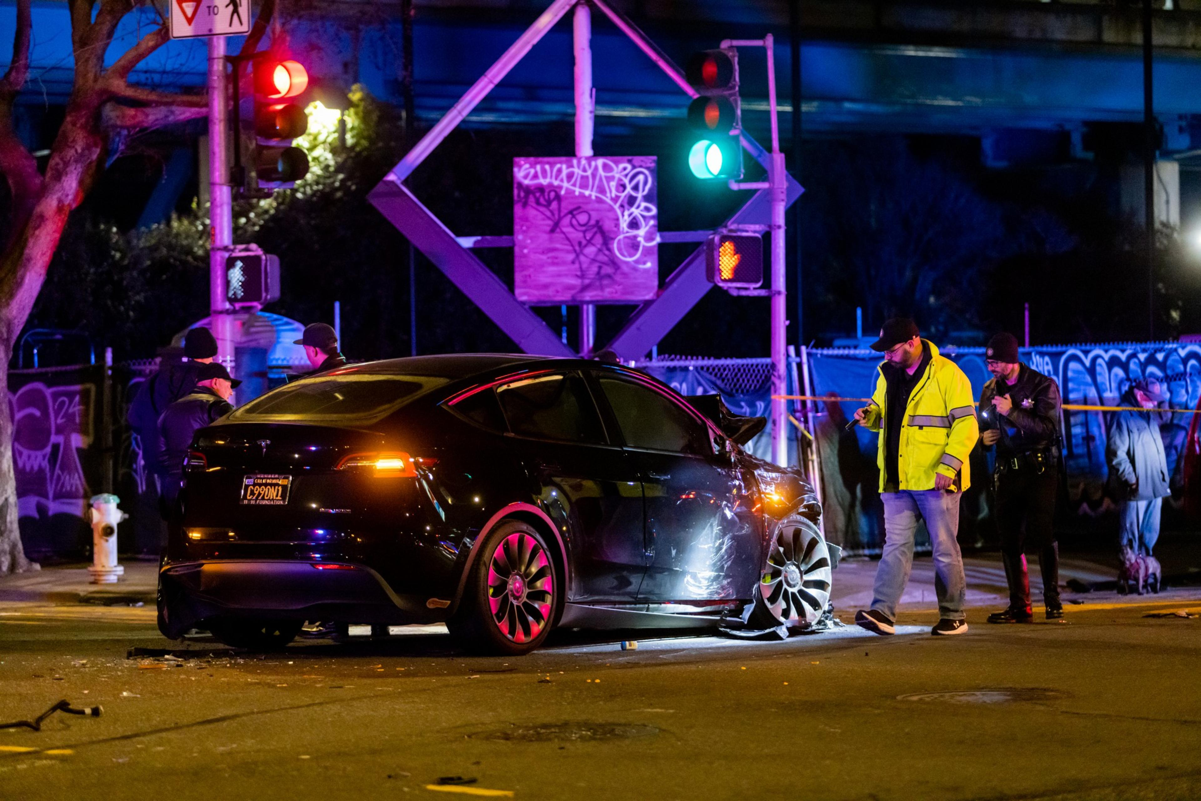 A damaged black car is at an intersection at night. People in reflective gear inspect the scene. Traffic lights are red and green, and there's graffiti on a nearby sign.